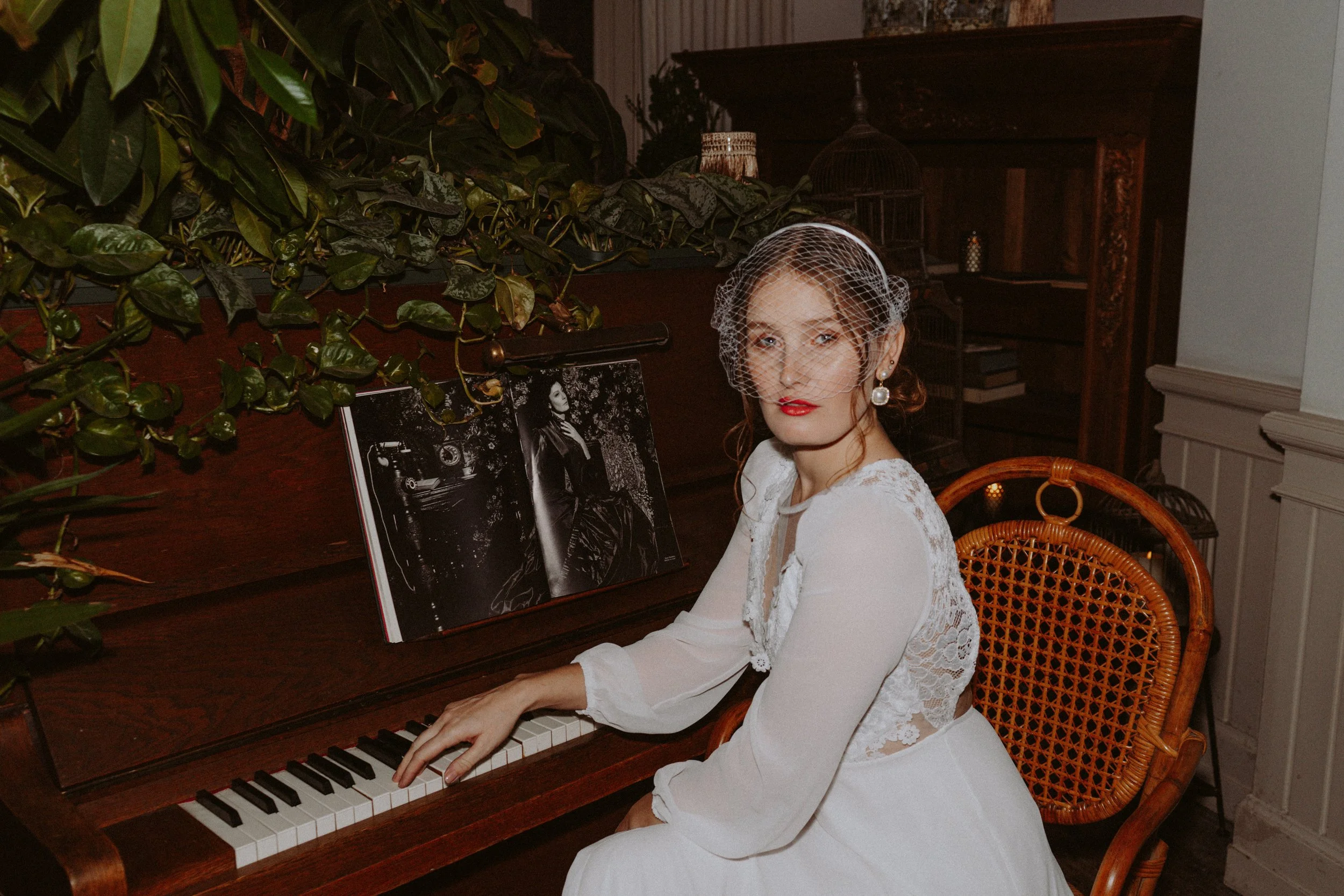 A woman with red hair, dressed in a white lace dress and pearl earrings, sitting at a piano with sheet music, surrounded by vintage decor and green plants.