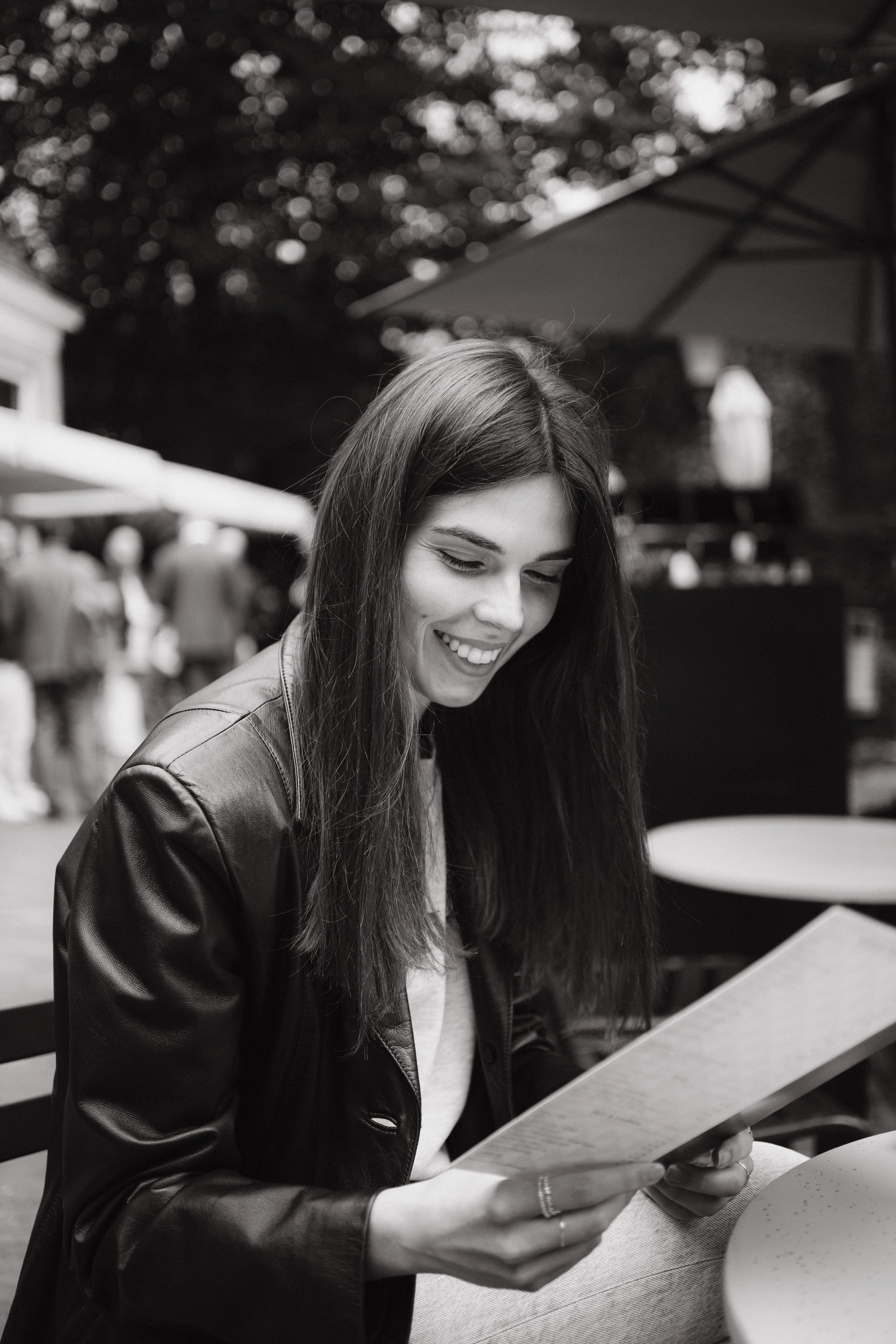 A young woman with long dark hair, smiling and reading a menu at a table in an outdoor setting, with a large umbrella and blurred background of other people and trees.