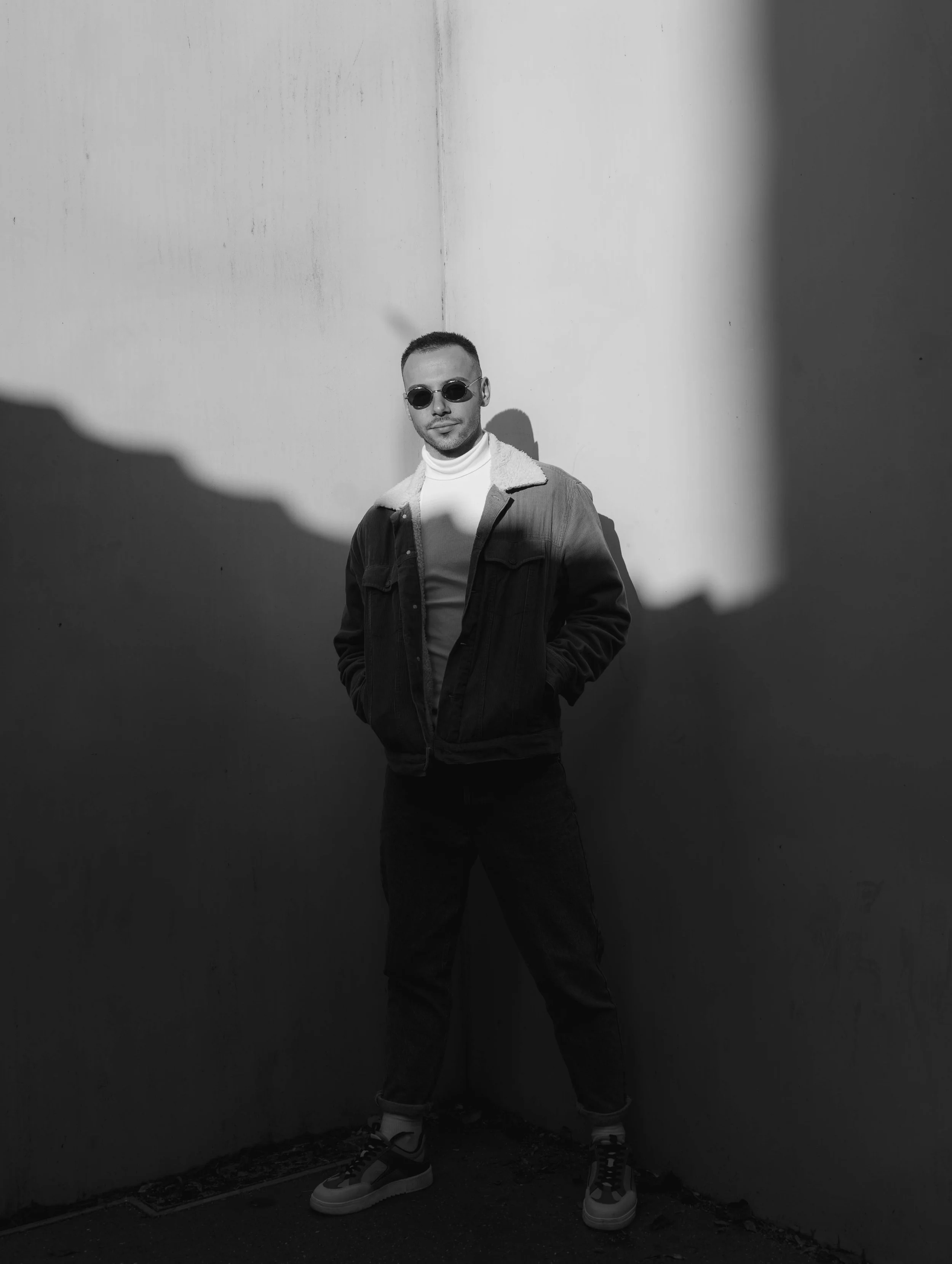 A man in sunglasses and a jacket standing against a plain wall with shadows, in black and white.