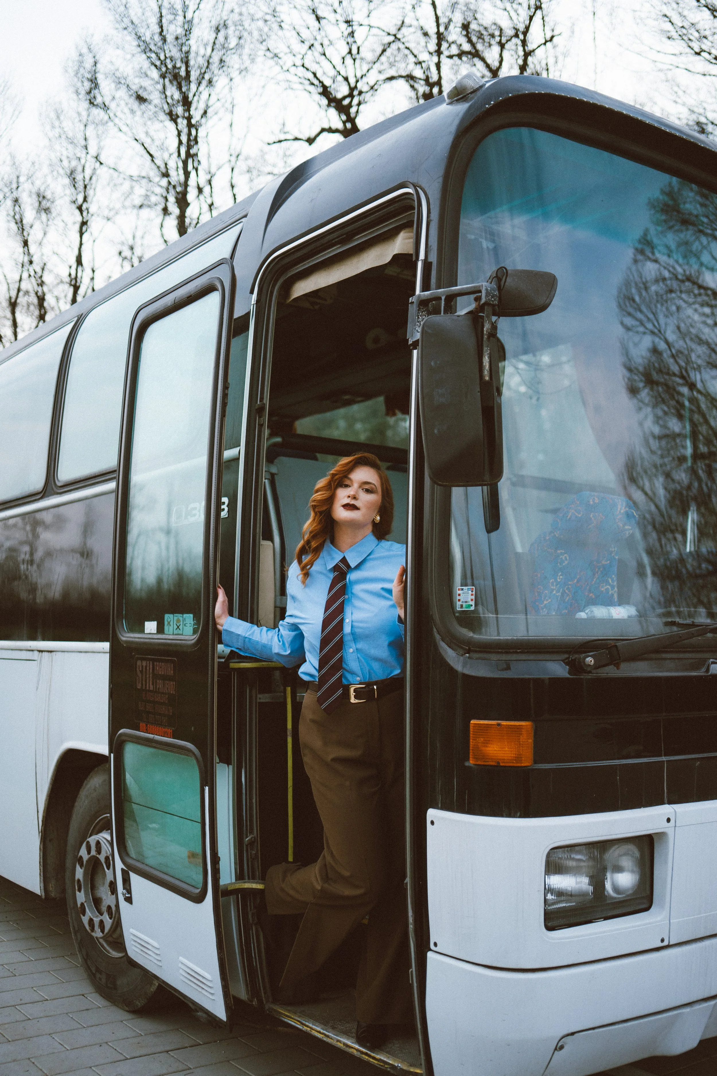 A woman with red hair, wearing a blue shirt and a tie, standing in the entrance of a large coach bus, holding the door open.