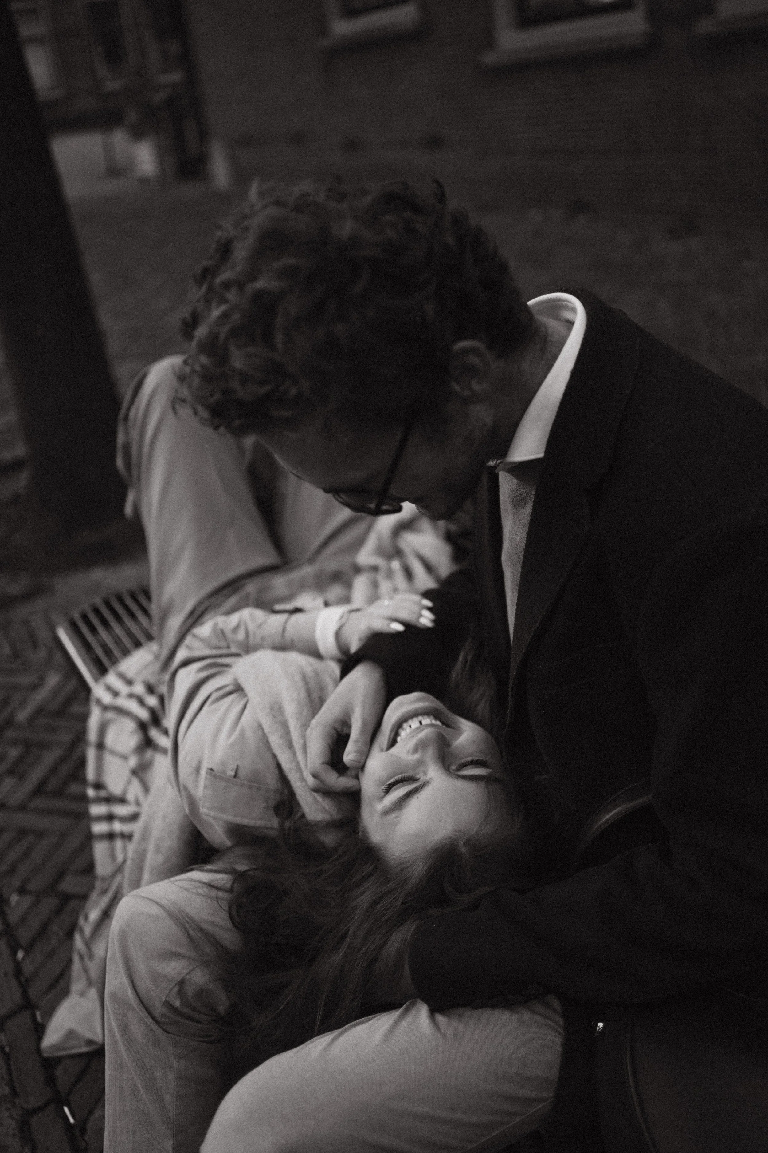 A man and woman share a joyful moment, with the woman lying back and smiling, and the man gently leaning over her. They are outdoors, in a casual setting.