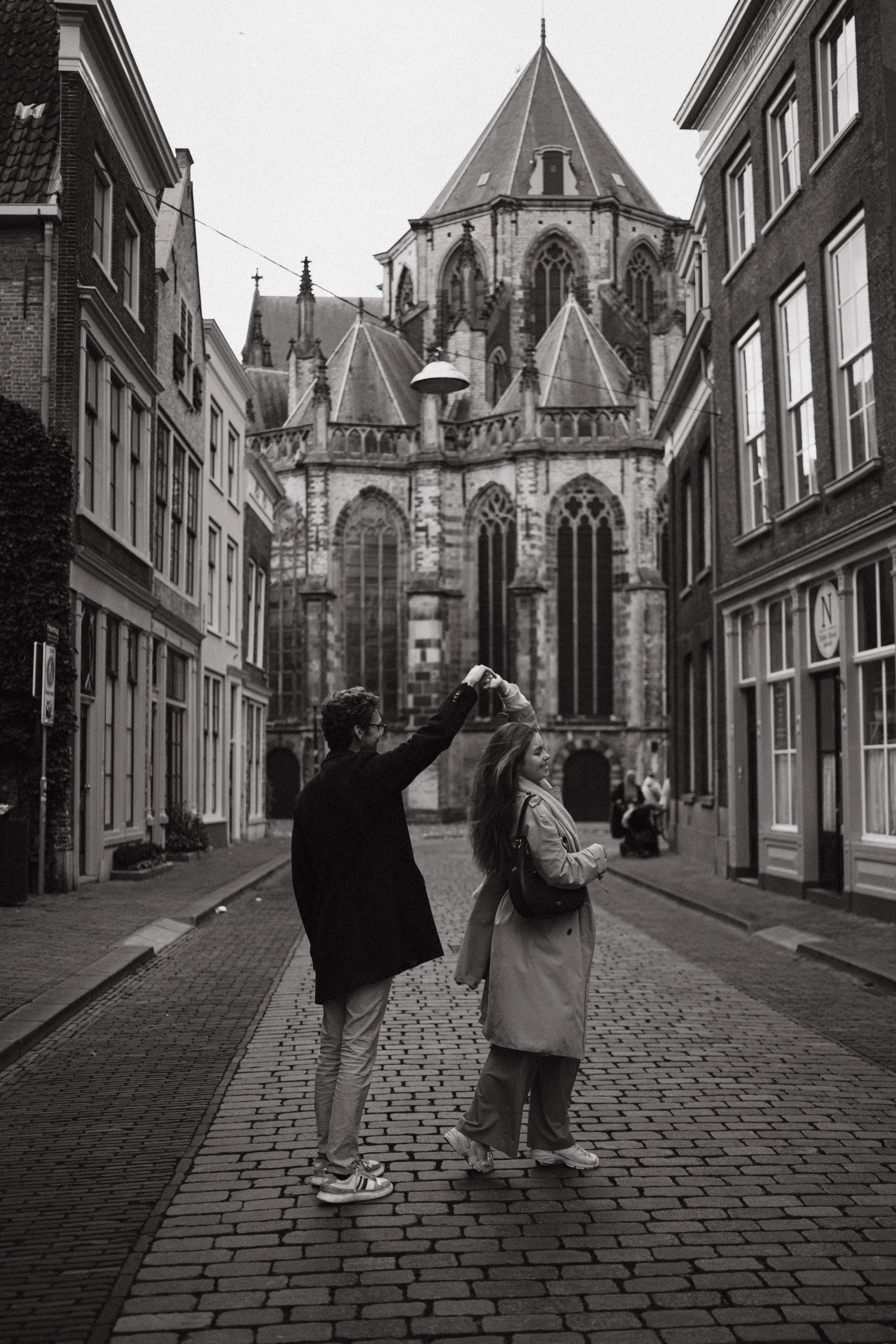 A black-and-white photo of a man and a woman standing on a cobblestone street in front of a large Gothic-style church. The man is holding up the woman's hand as if they are dancing or sharing a moment. The woman is looking up at the man, smiling. The street is lined with old buildings, and there are streetlights and other people in the background.