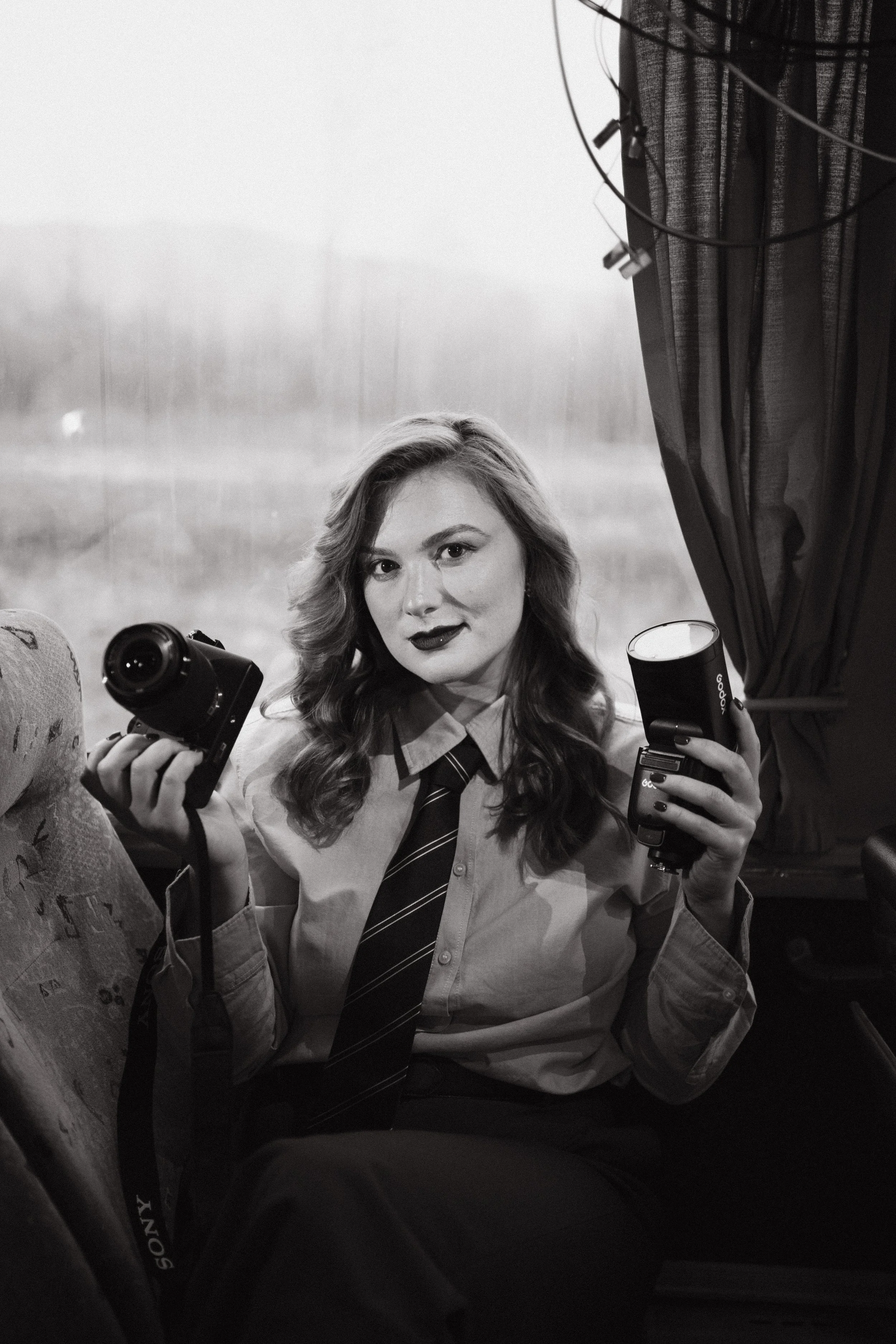 Black and white photo of a young woman in a school uniform holding a camera and a microphone, sitting inside a vehicle with a window showing a blurred outdoor landscape.