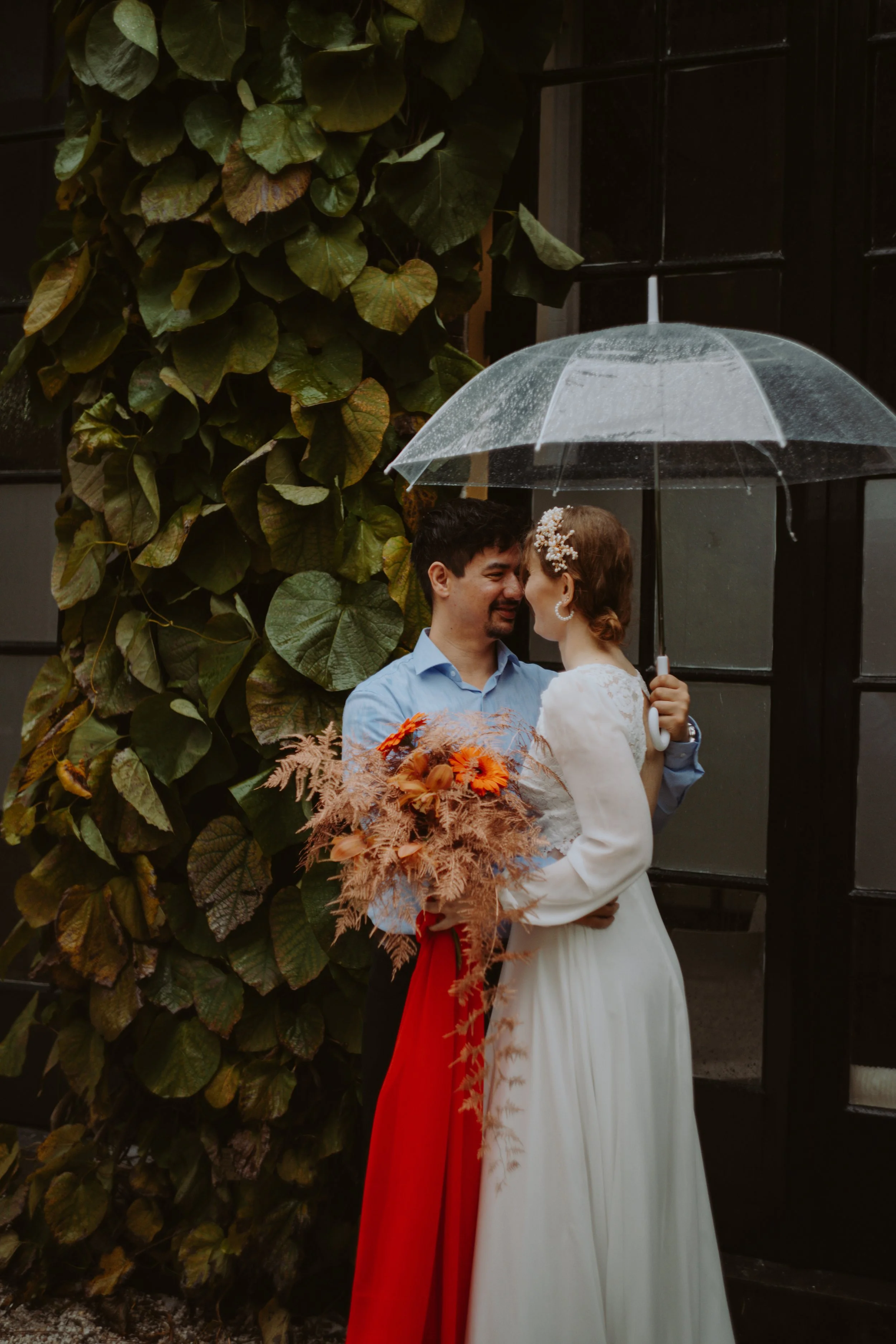 A couple stands under a clear umbrella with raindrops, sharing a kiss in front of a lush wall of green leaves, with the woman holding a bouquet of orange and peach flowers.