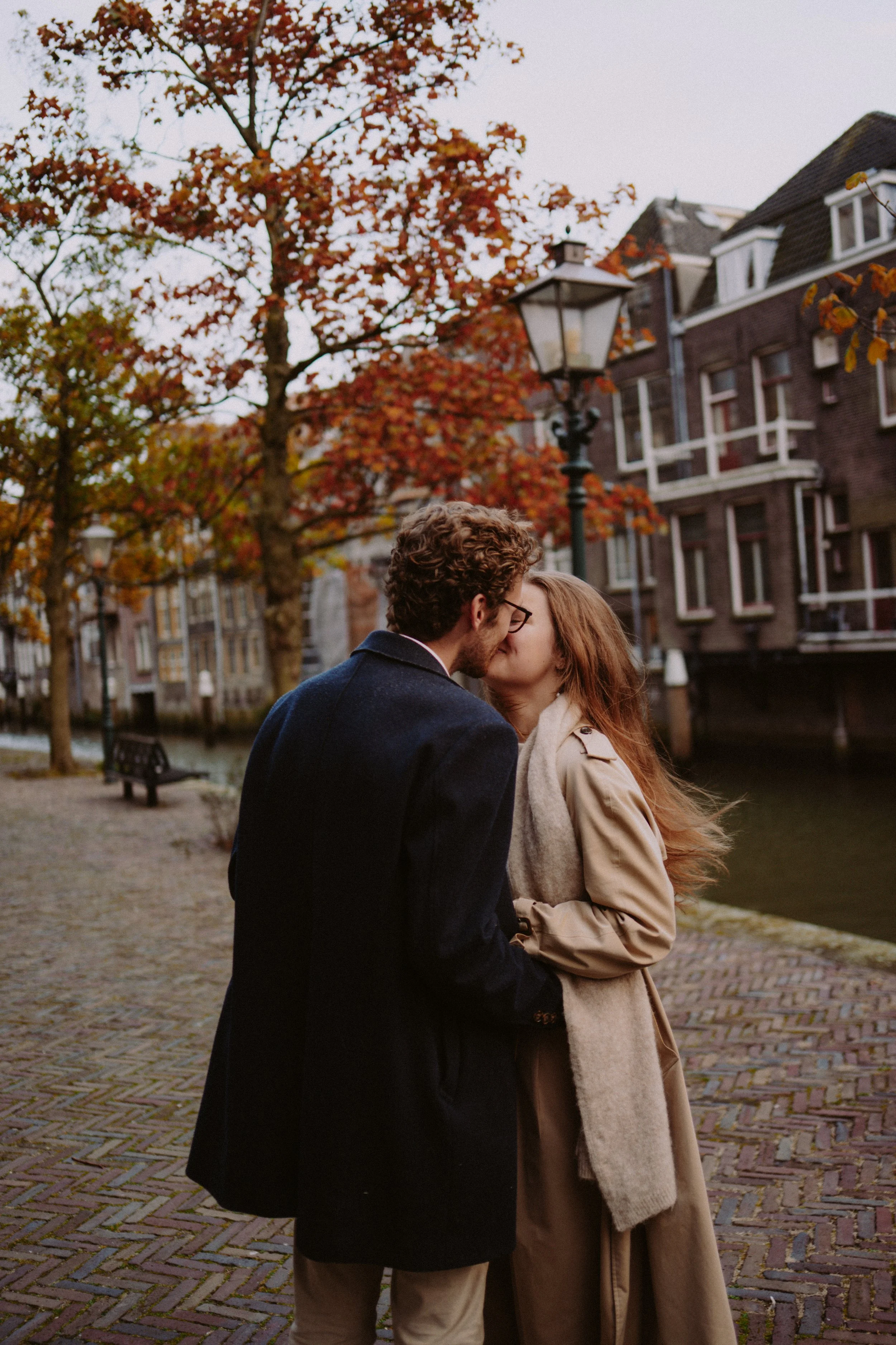 A couple is kissing outdoors on a cobblestone path, surrounded by autumn trees with red and orange leaves, near a canal and historic brick buildings with gabled roofs.