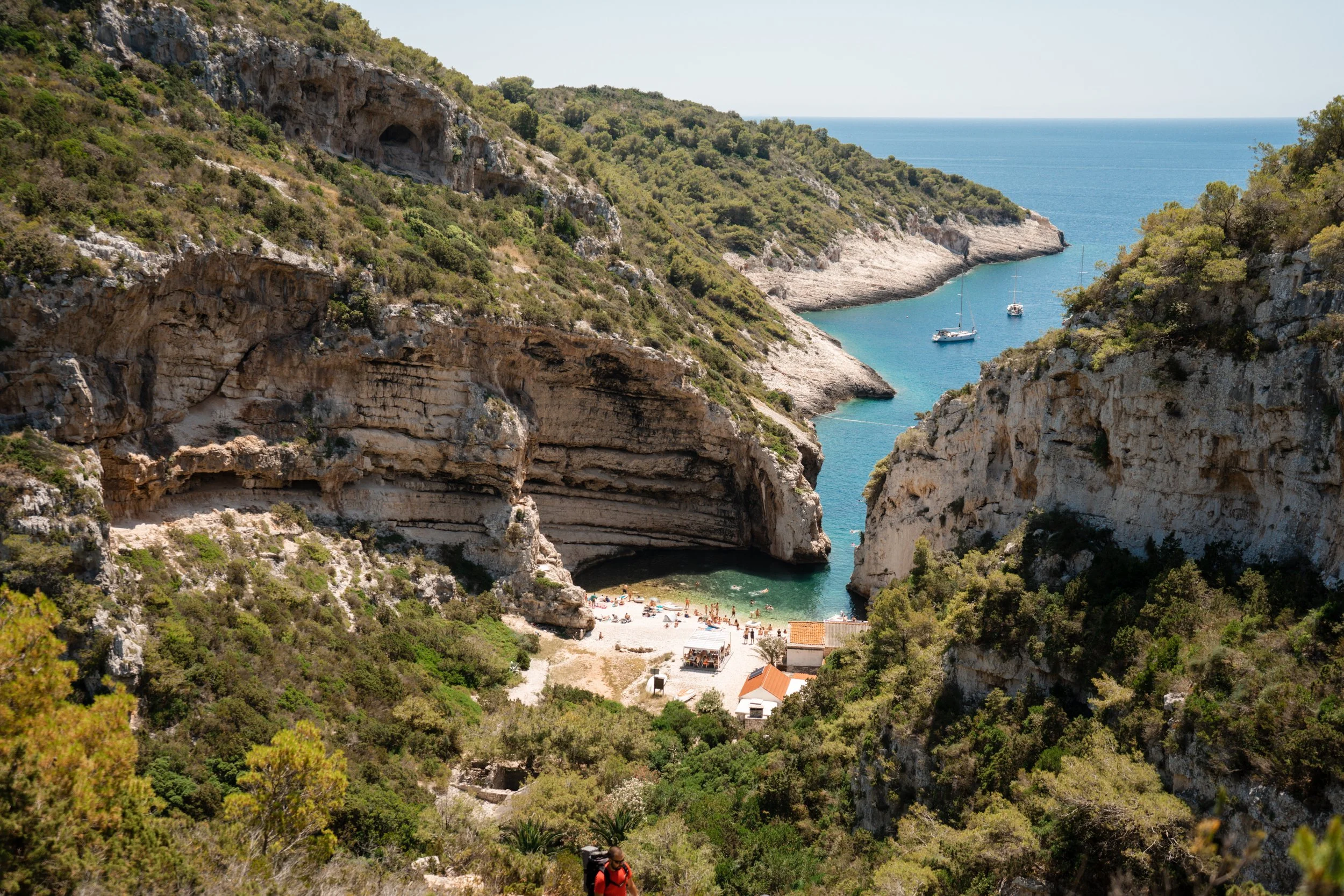 A coastal scene with steep, rocky hills covered in green foliage surrounding a small beach and turquoise water bay with sailboats. A few people are visible at the beach, and a hiker in a red shirt is in the foreground.