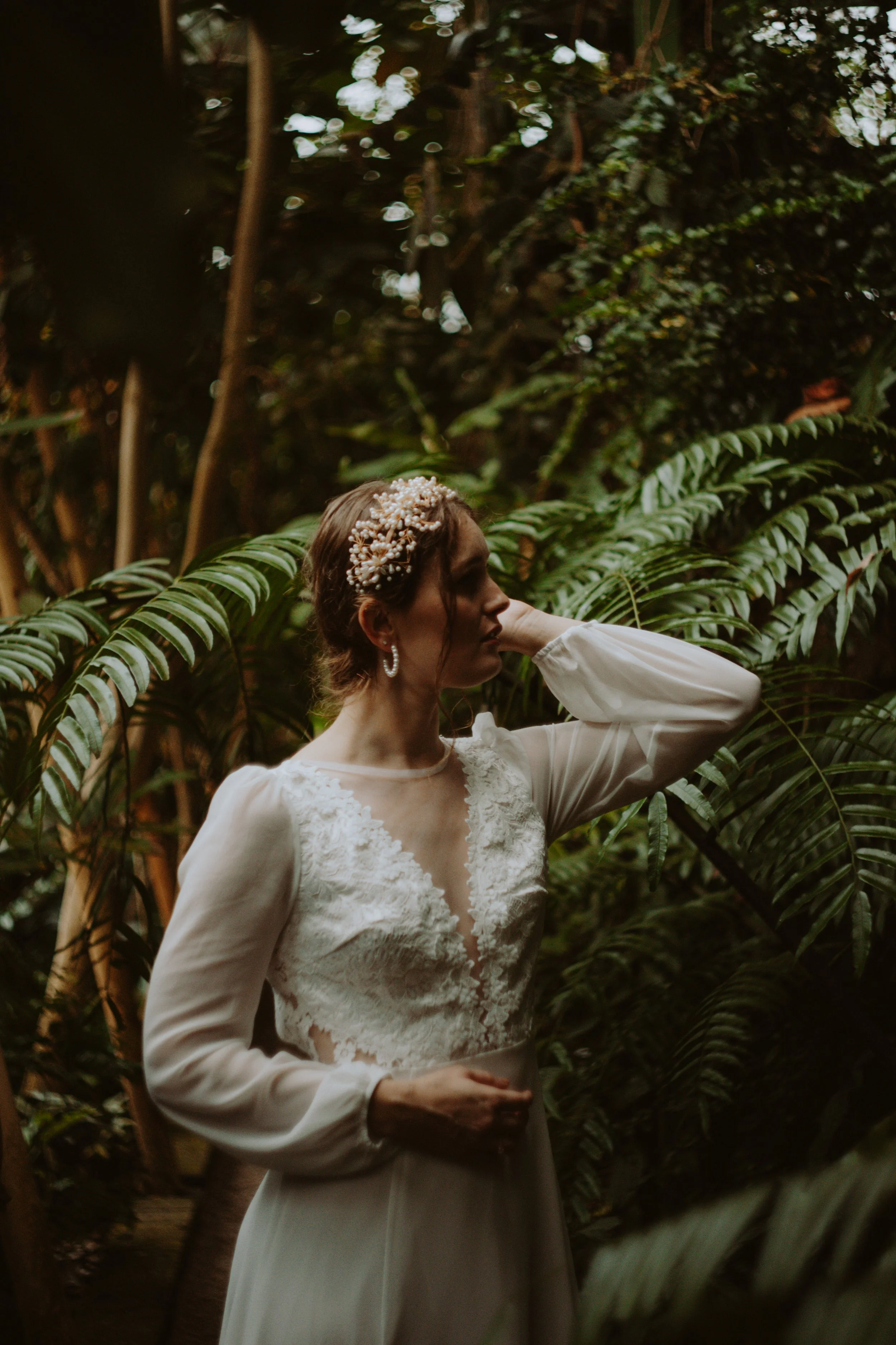 A woman in a white dress with lace details, standing among green tropical plants, wearing a pearl headpiece and hoop earrings, with one hand near her face and the other on her waist, in a lush outdoor setting.