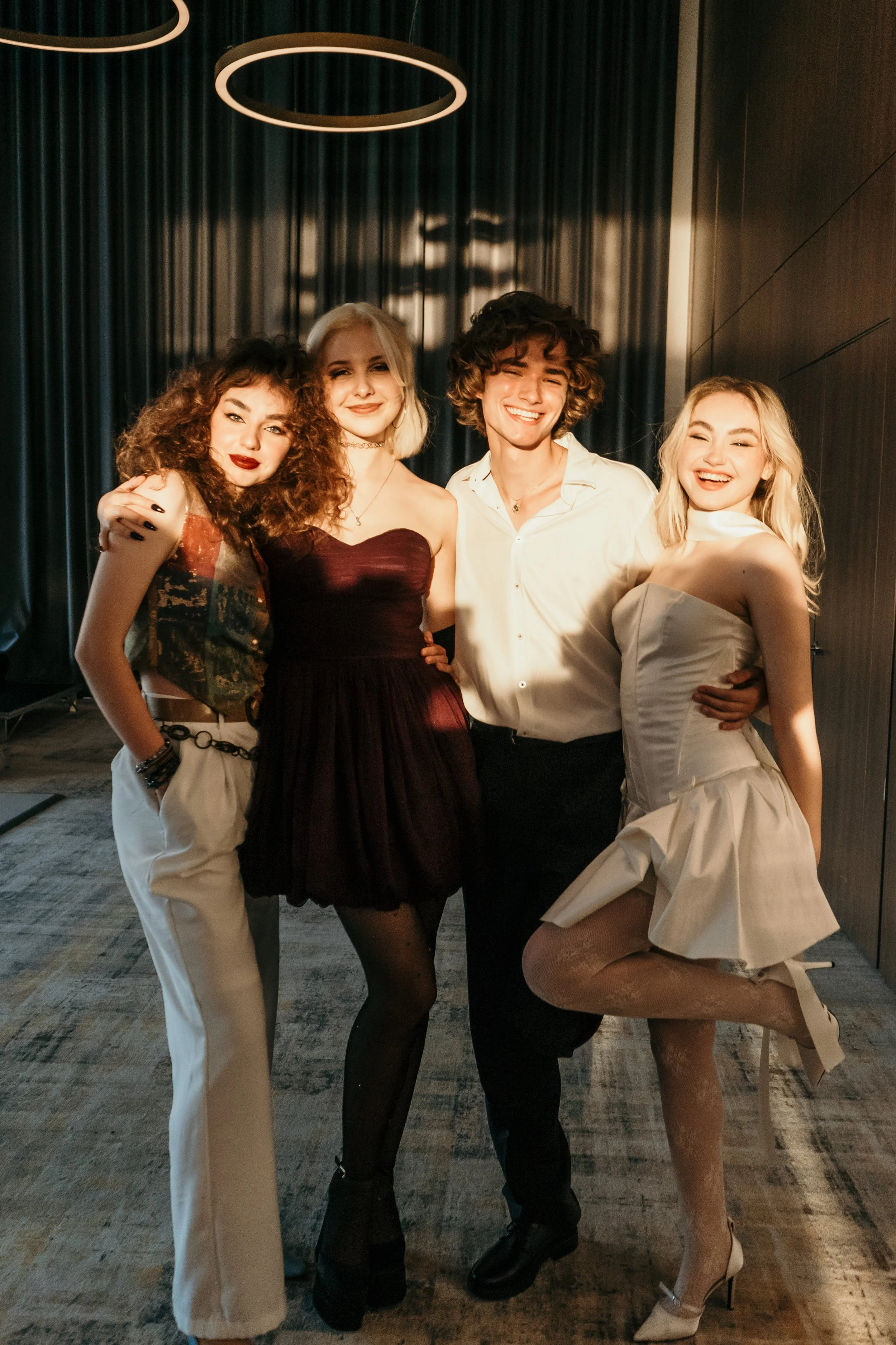 Four young adults celebrating together, dressed in fashionable attire, smiling, and posing with their arms around each other in a dimly lit indoor setting.