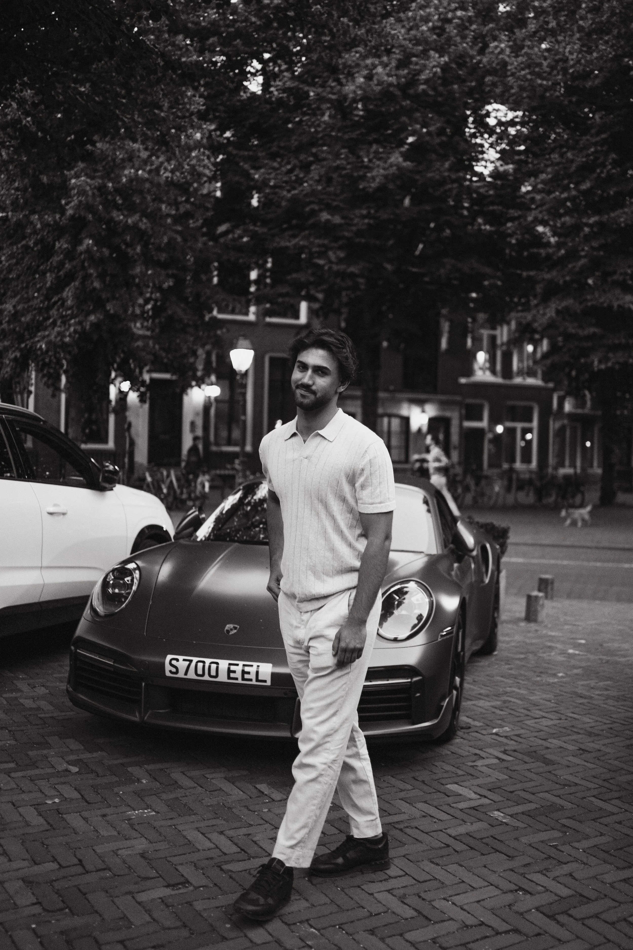 A young man with dark hair and a beard walking on a city street at night, wearing a light-colored polo shirt and white pants, with parked cars and trees in the background.