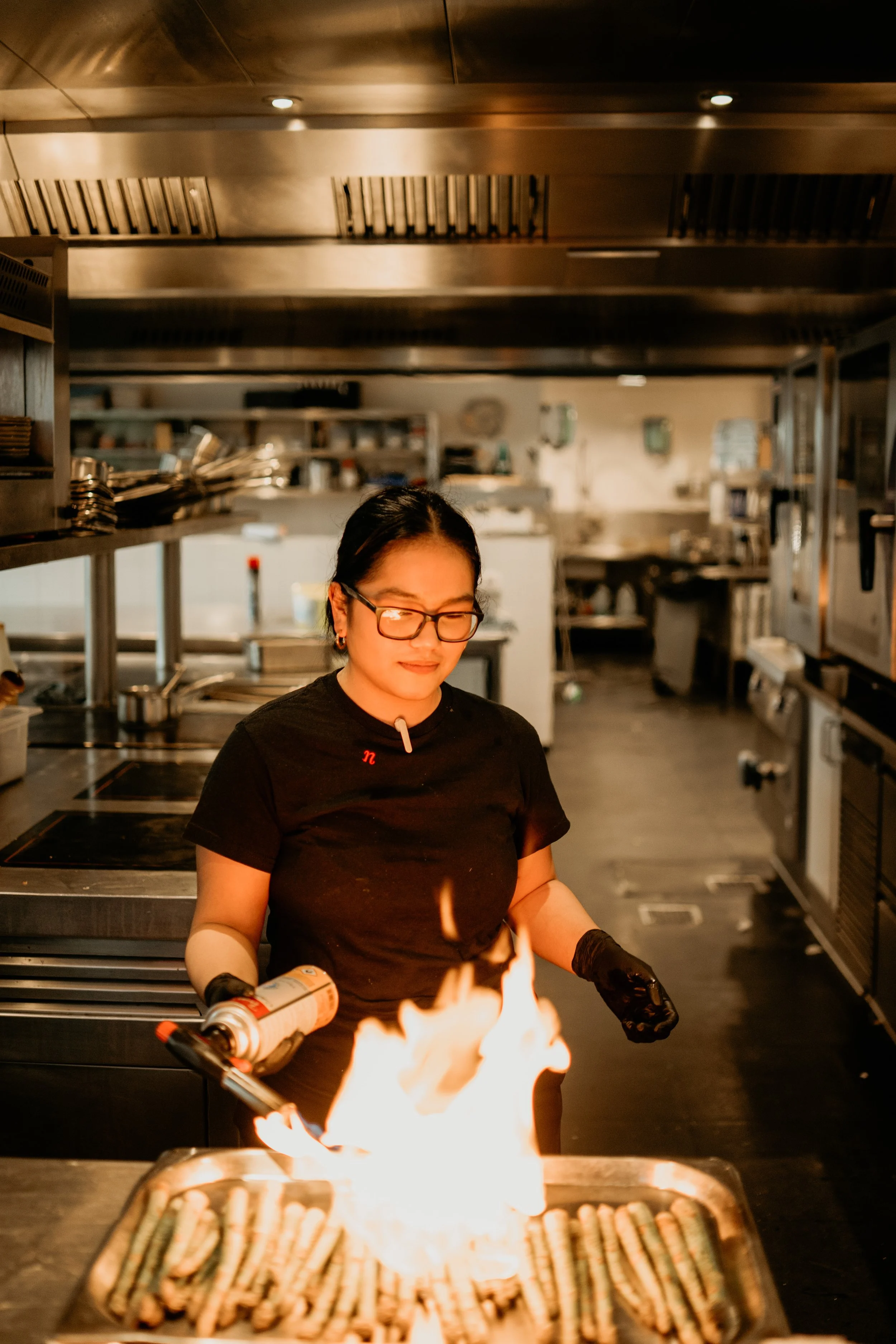 A woman cooking over a grill with flames in a commercial kitchen, holding a can of cooking spray in one hand and a utensil in the other.