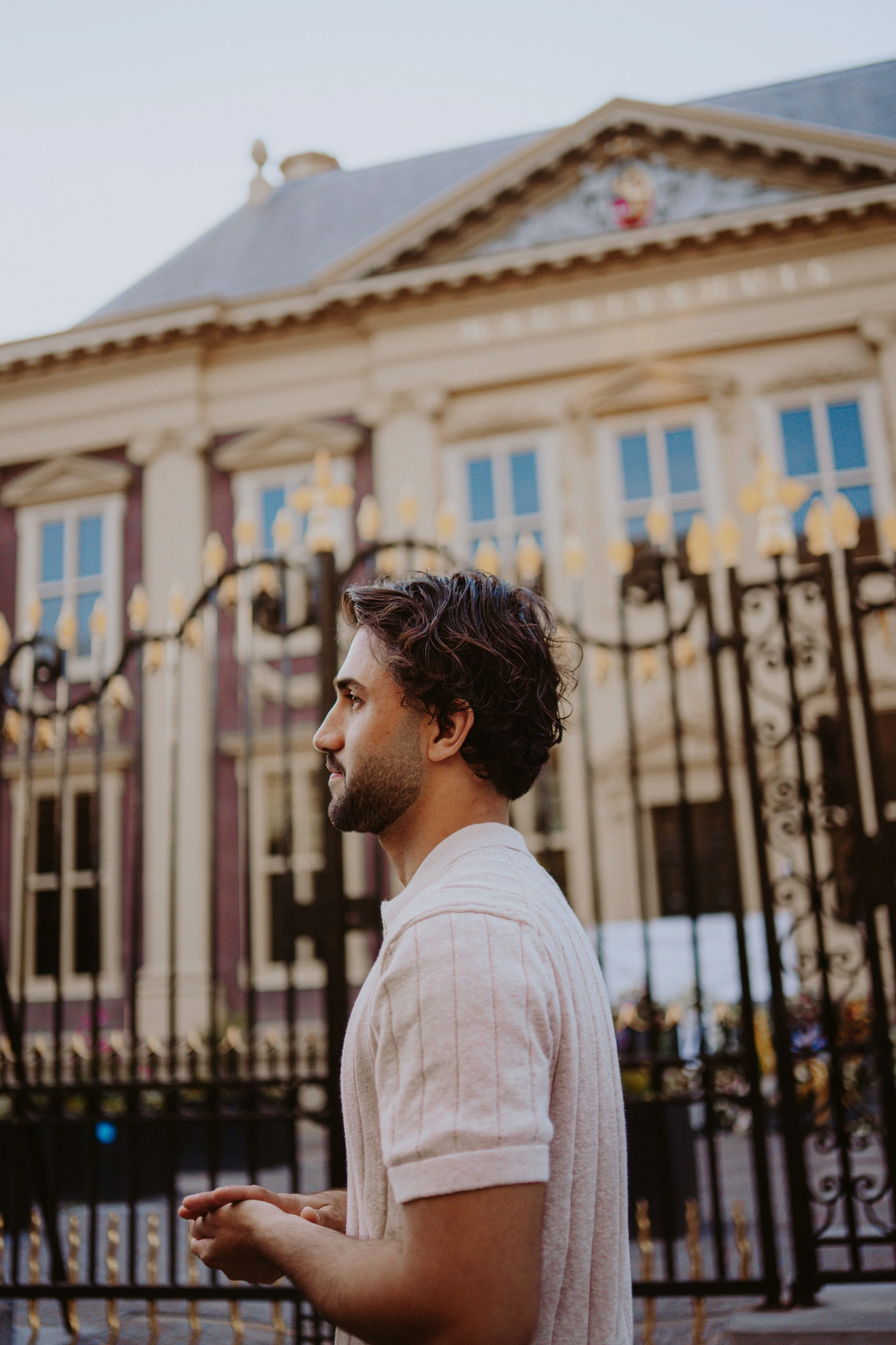 A young man with dark wavy hair and a beard, wearing a light-colored v-neck shirt, standing in profile in front of a decorative wrought iron fence with a building in the background.