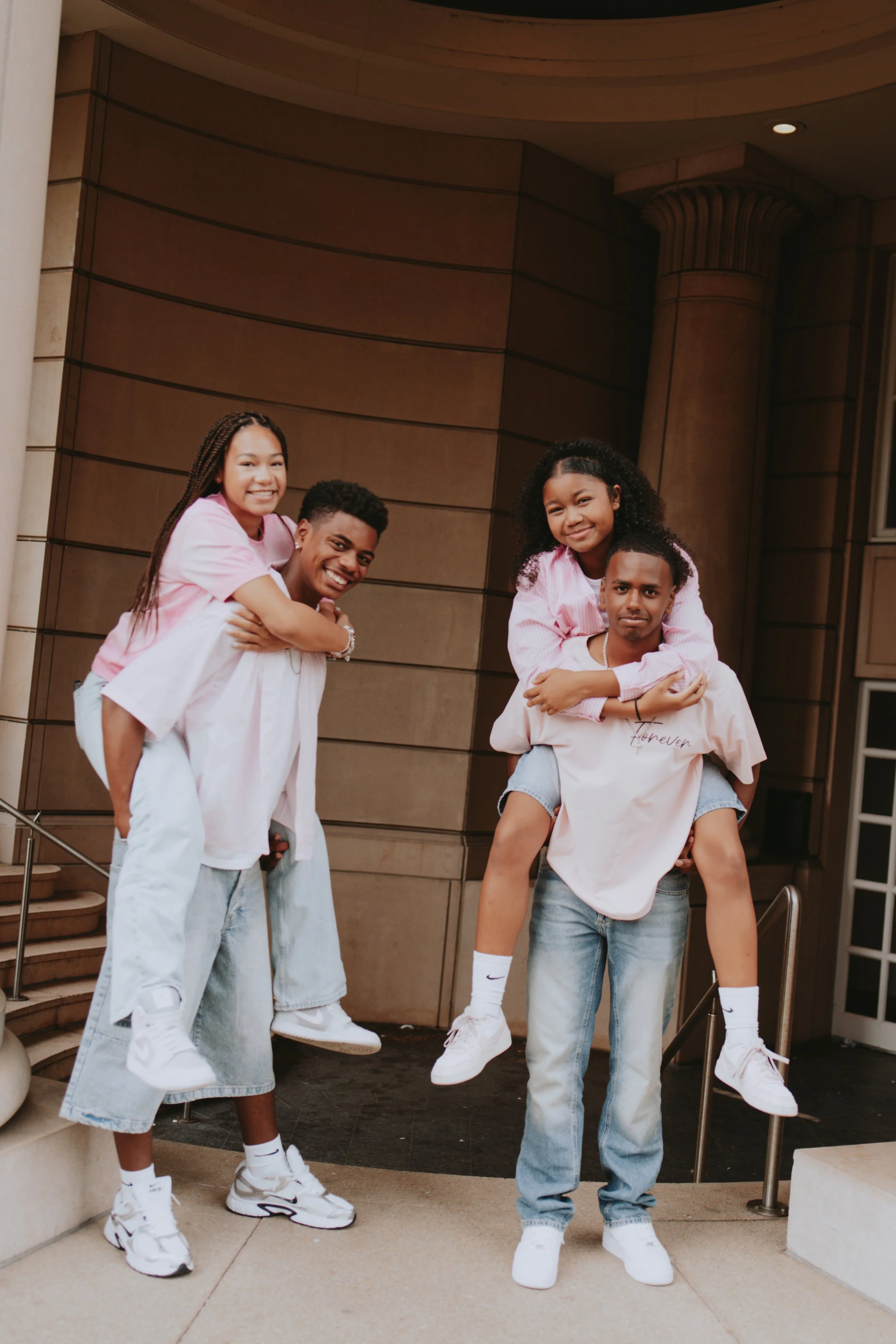 Group of five young people smiling and posing outside a building, some giving piggyback rides, wearing casual clothing including pink and white shirts, jeans, and sneakers.
