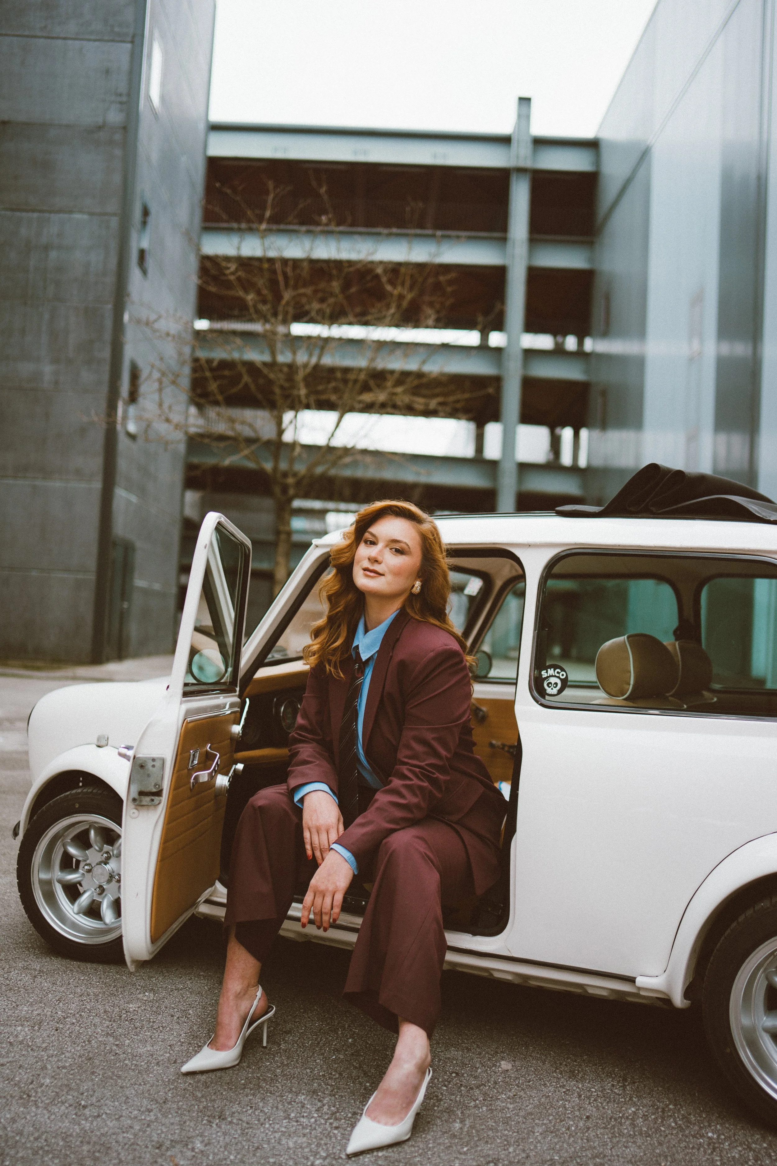 Personal branding photo of a business woman with long, wavy hair and wearing high heels and a brown suit is sitting with one leg crossed outside a vintage white car. The car door is open, and she is looking at the camera with a confident expression. 