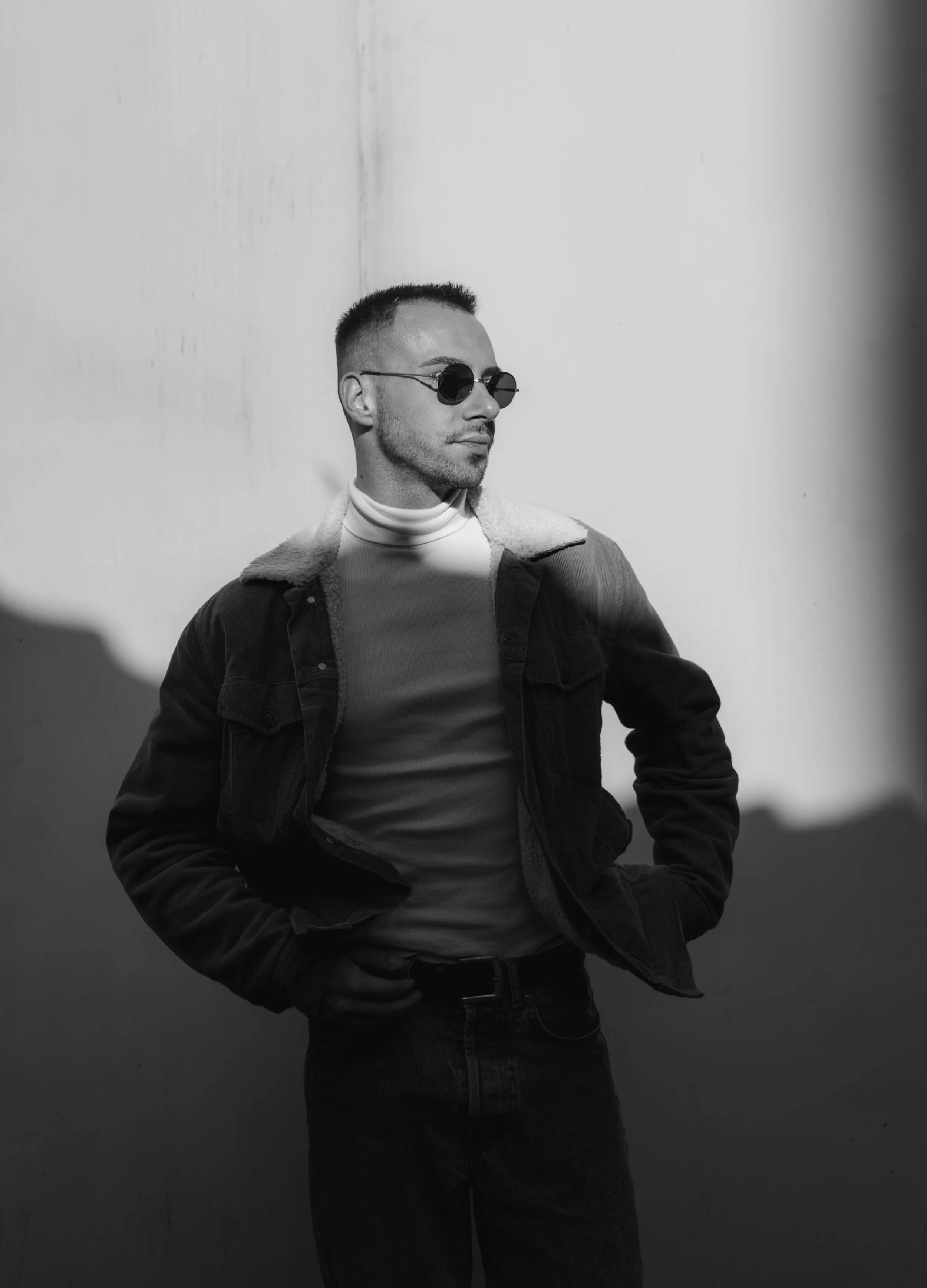A young man with short hair, wearing round sunglasses, a turtleneck, and a jacket with a shearling collar, posing against a plain wall in black and white.