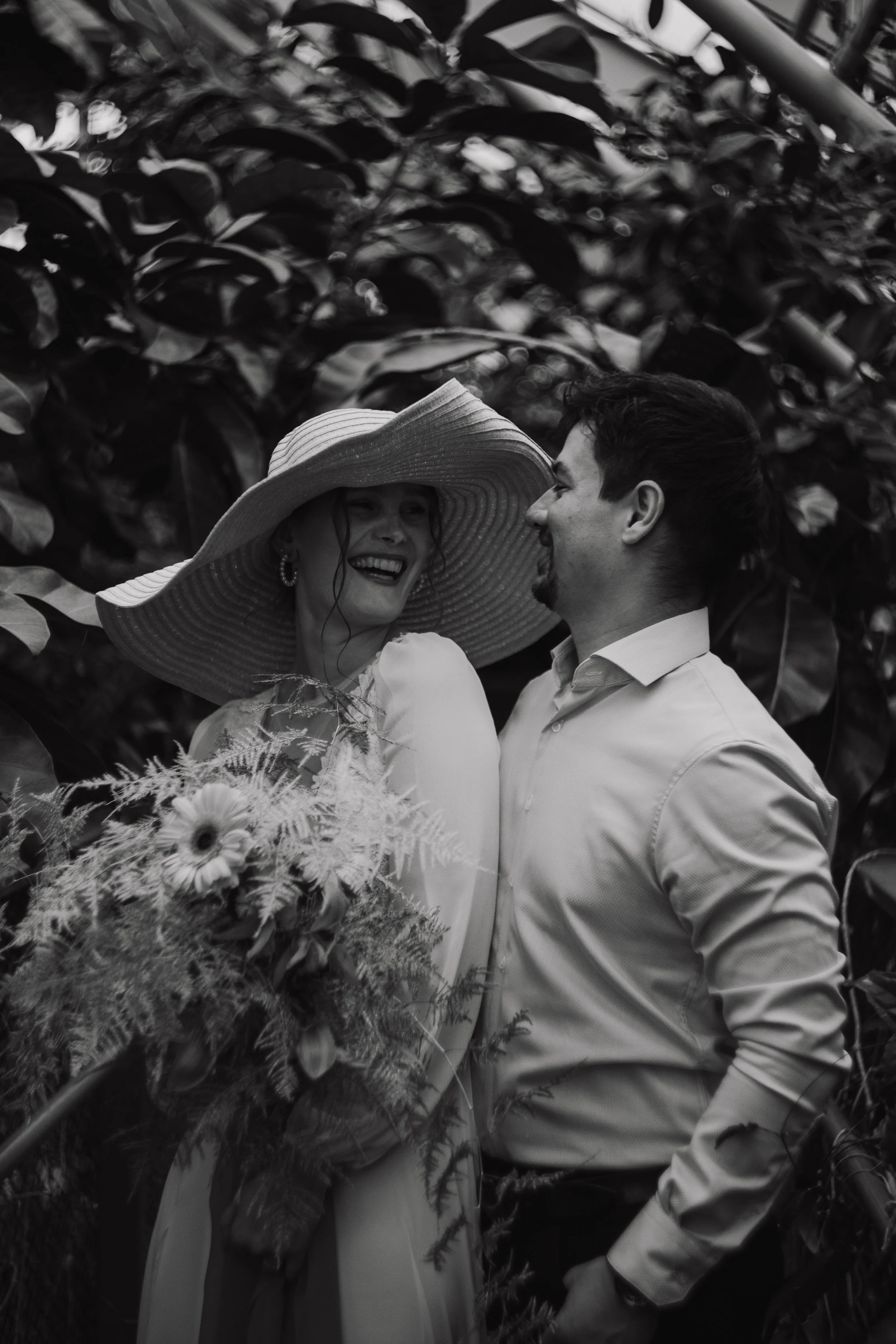 A man and woman smile and look at each other, with the woman wearing a large hat and holding a bouquet of flowers. They are standing outdoors surrounded by dense foliage.