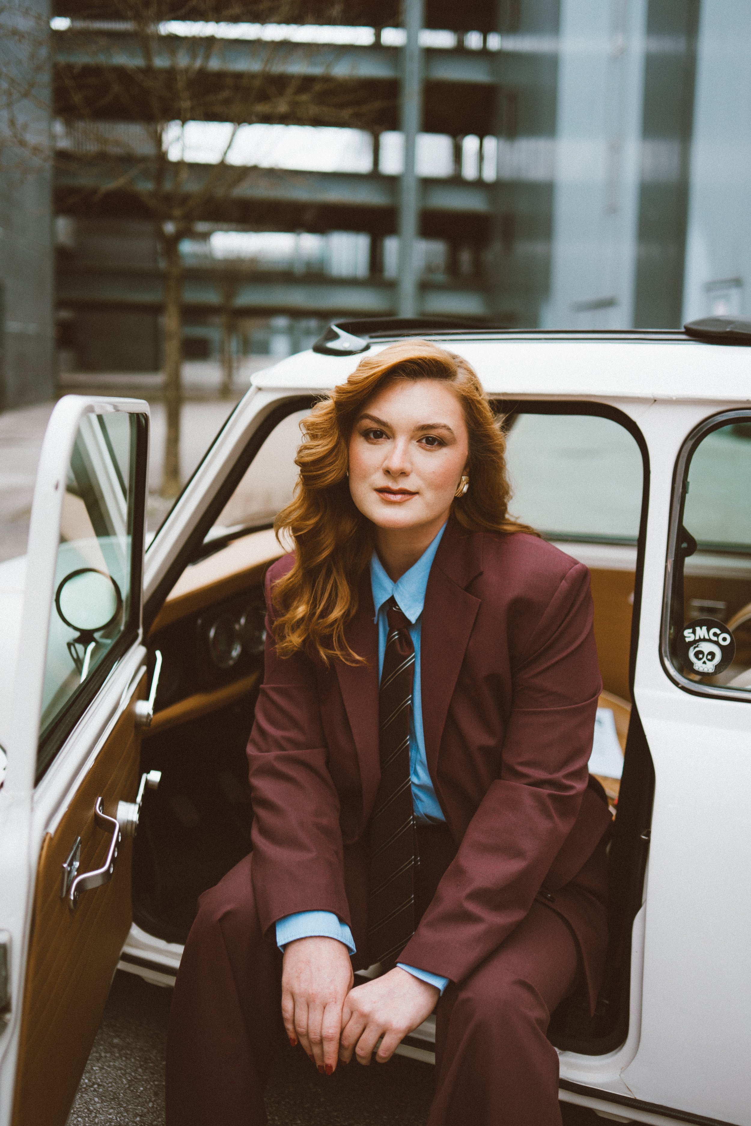 A woman with red hair, wearing a maroon suit with a blue shirt and tie, sitting in a vintage white car with the door open, in an urban environment with buildings and trees in the background.