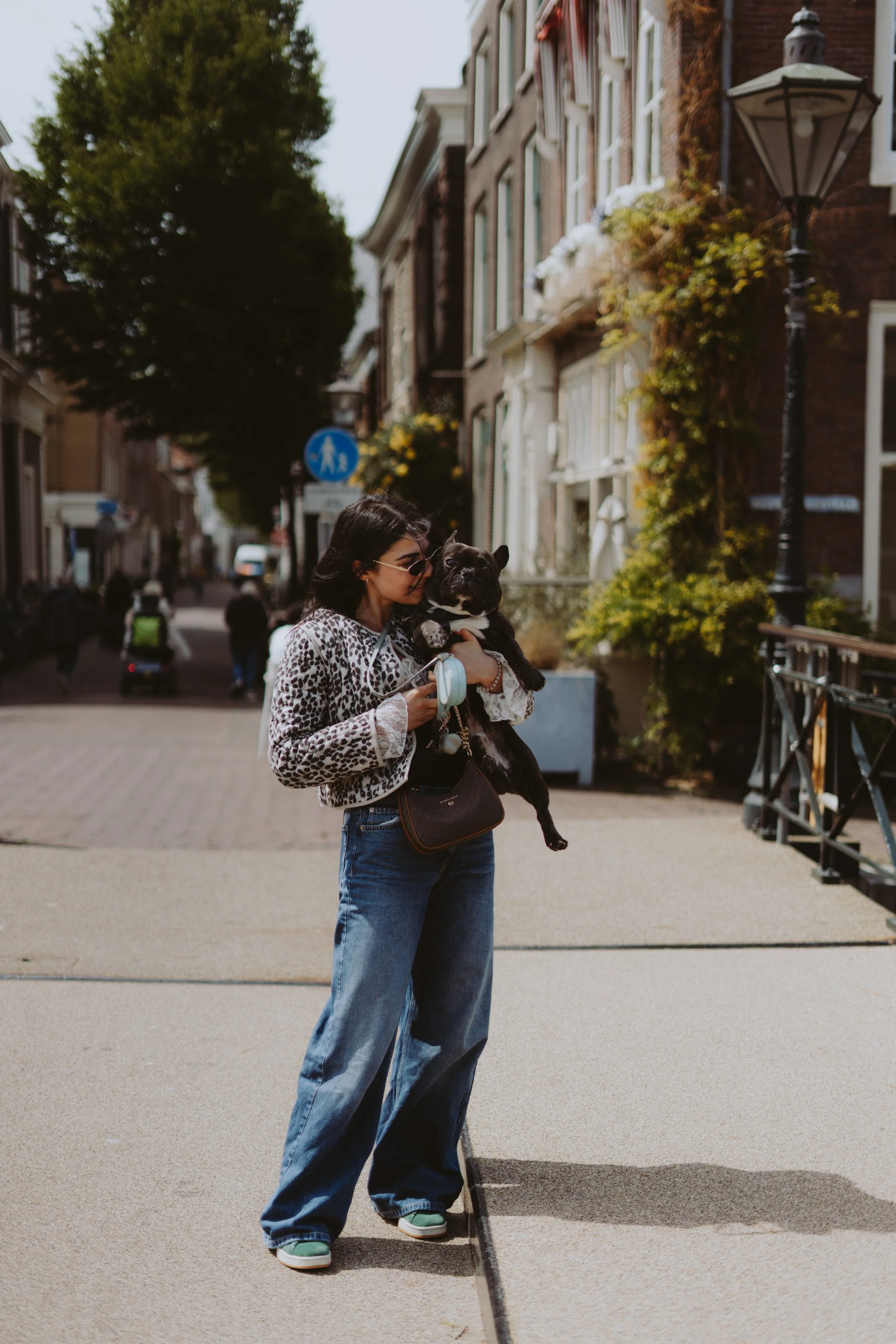 A woman with dark hair, wearing sunglasses, a leopard print jacket, and high-waisted jeans, holding a small black dog, walking on a city sidewalk with buildings, trees, and street lamps in the background.