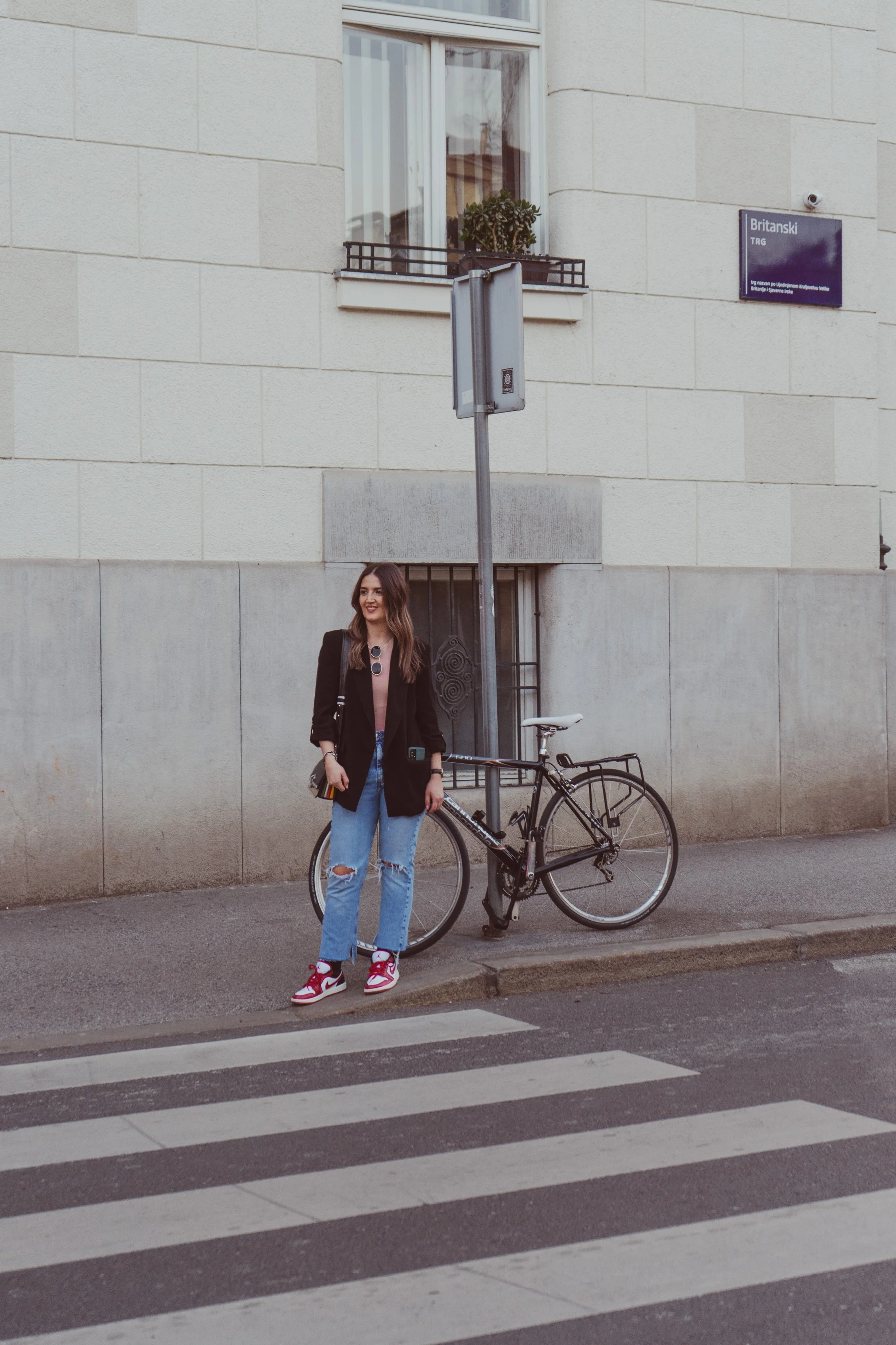 A woman with long brown hair, wearing a black blazer, ripped jeans, and red sneakers, stands on a city street next to a bicycle. She is smiling and holding a small black purse. Behind her is a concrete building with one window, a sign that says 'Brit