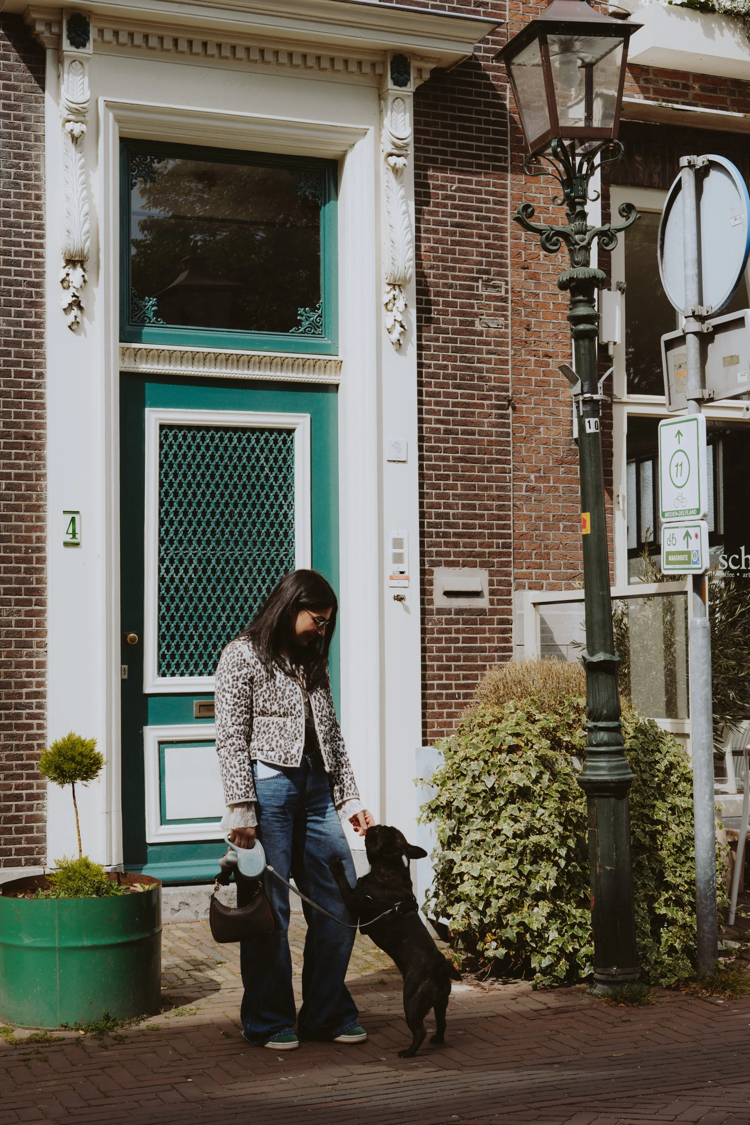 Woman with glasses wearing a leopard print jacket and blue jeans, holding a leash with a black dog, standing on a brick sidewalk in front of a teal and white door with intricate trim, near a large shrub and a green plant in a large pot, on a city str