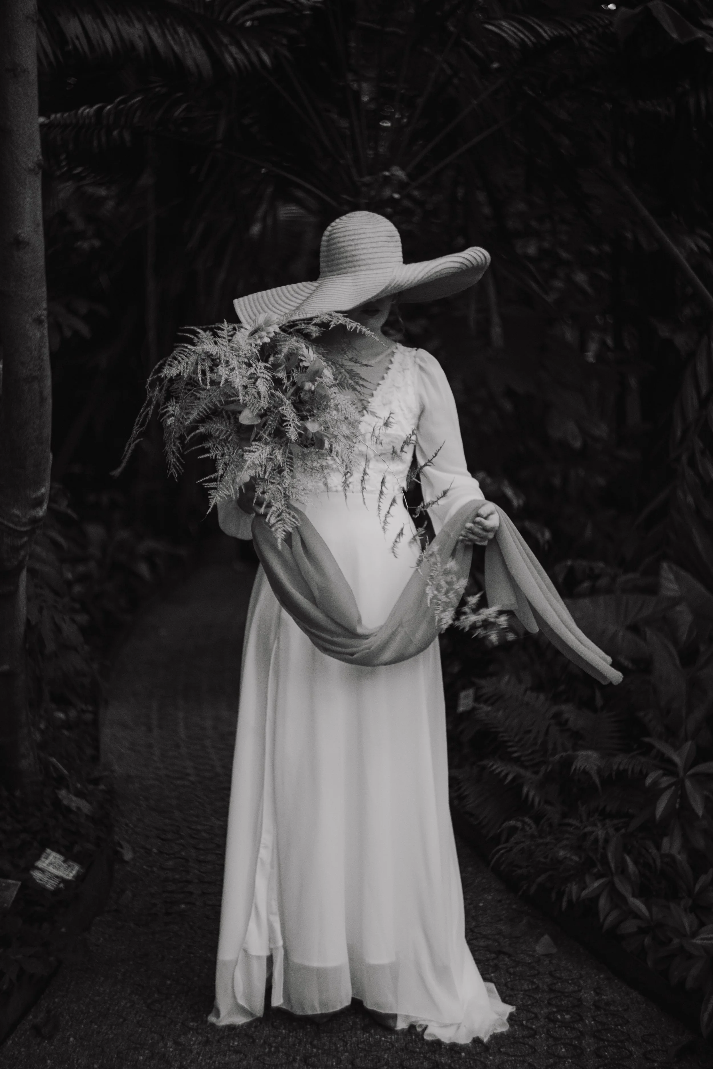A woman wearing a large straw hat and a long, flowing white dress, holding a bouquet of ferns, standing outdoors among lush greenery.