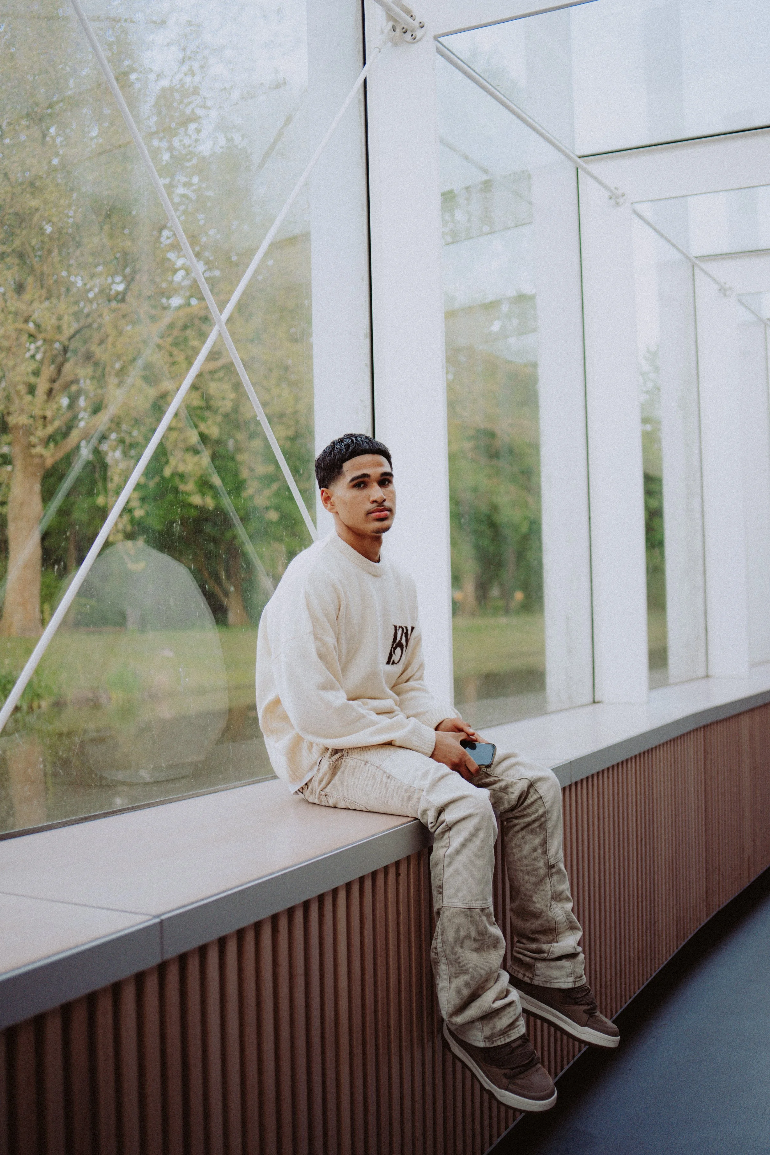 A young man with short dark hair and light brown skin is sitting on a bench near a large window in what appears to be a modern building or airport. He is wearing a cream-colored sweater, beige baggy pants, and brown and white sneakers. He is holding 
