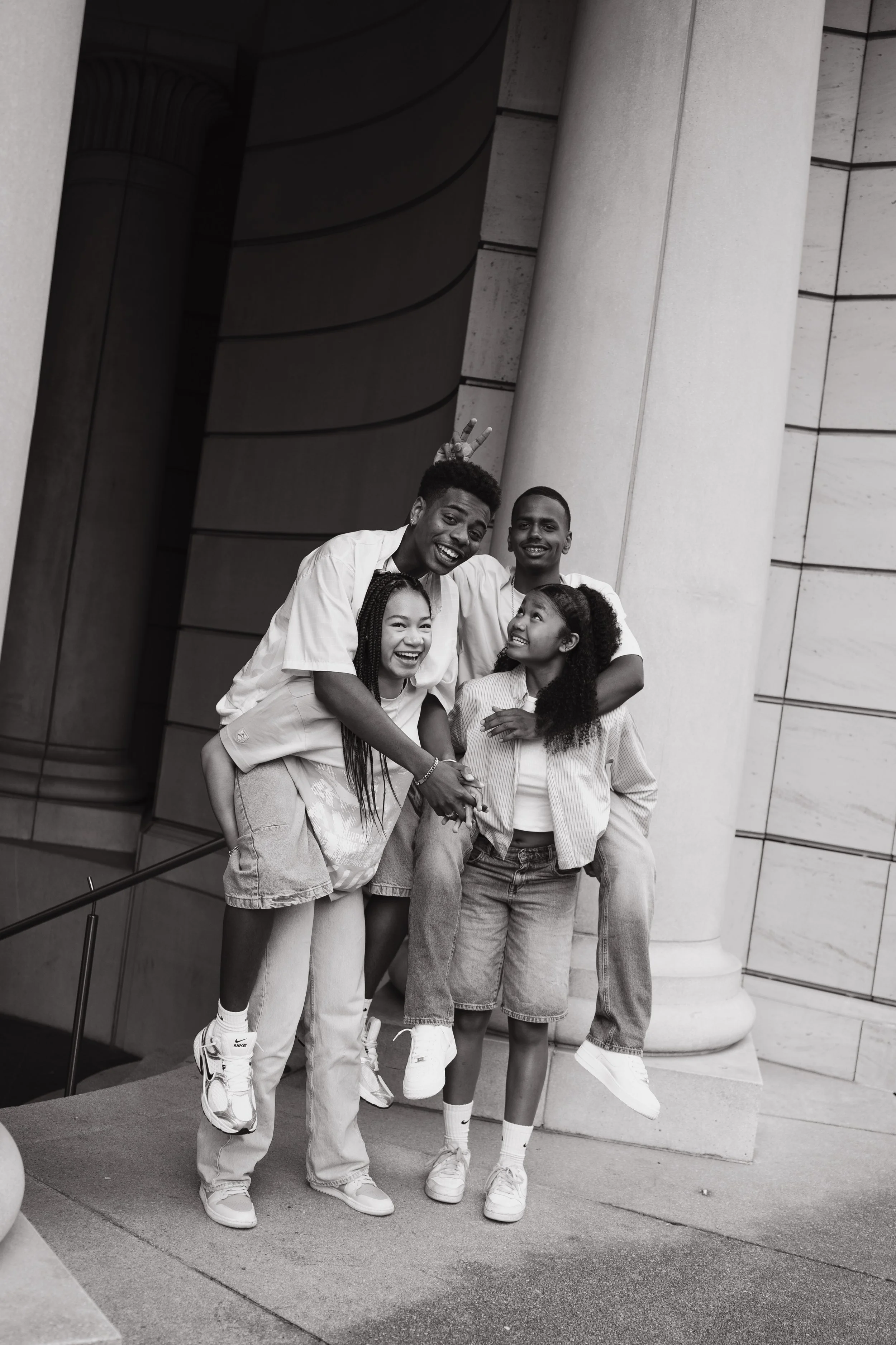 Four young people standing in front of a large stone column, posing for a photo. They appear happy and are making playful gestures, with one person giving bunny ears to another.