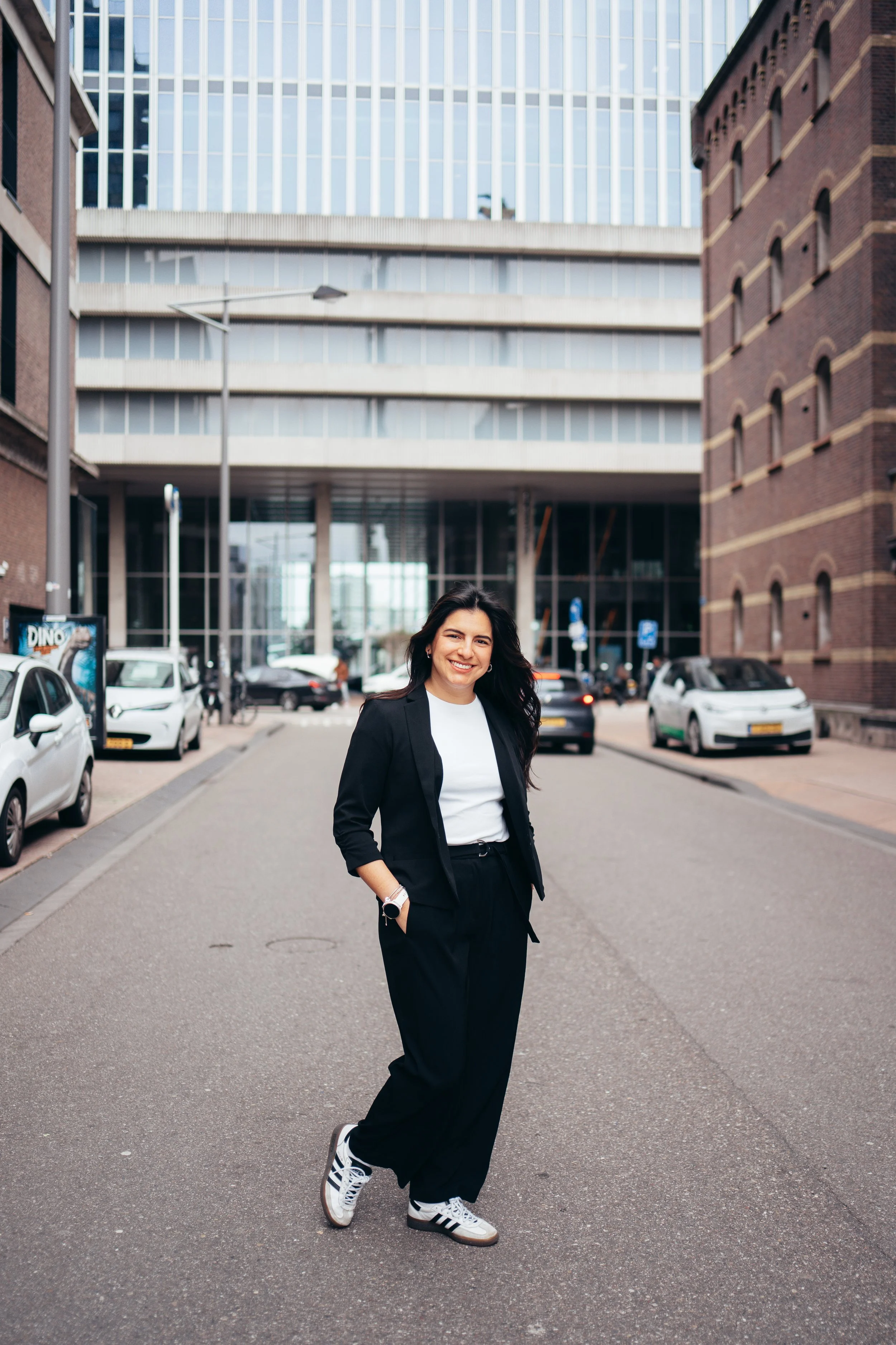 a woman in black pants, a white shirt, and a black blazer smiling on a city street with parked cars and modern buildings in the background