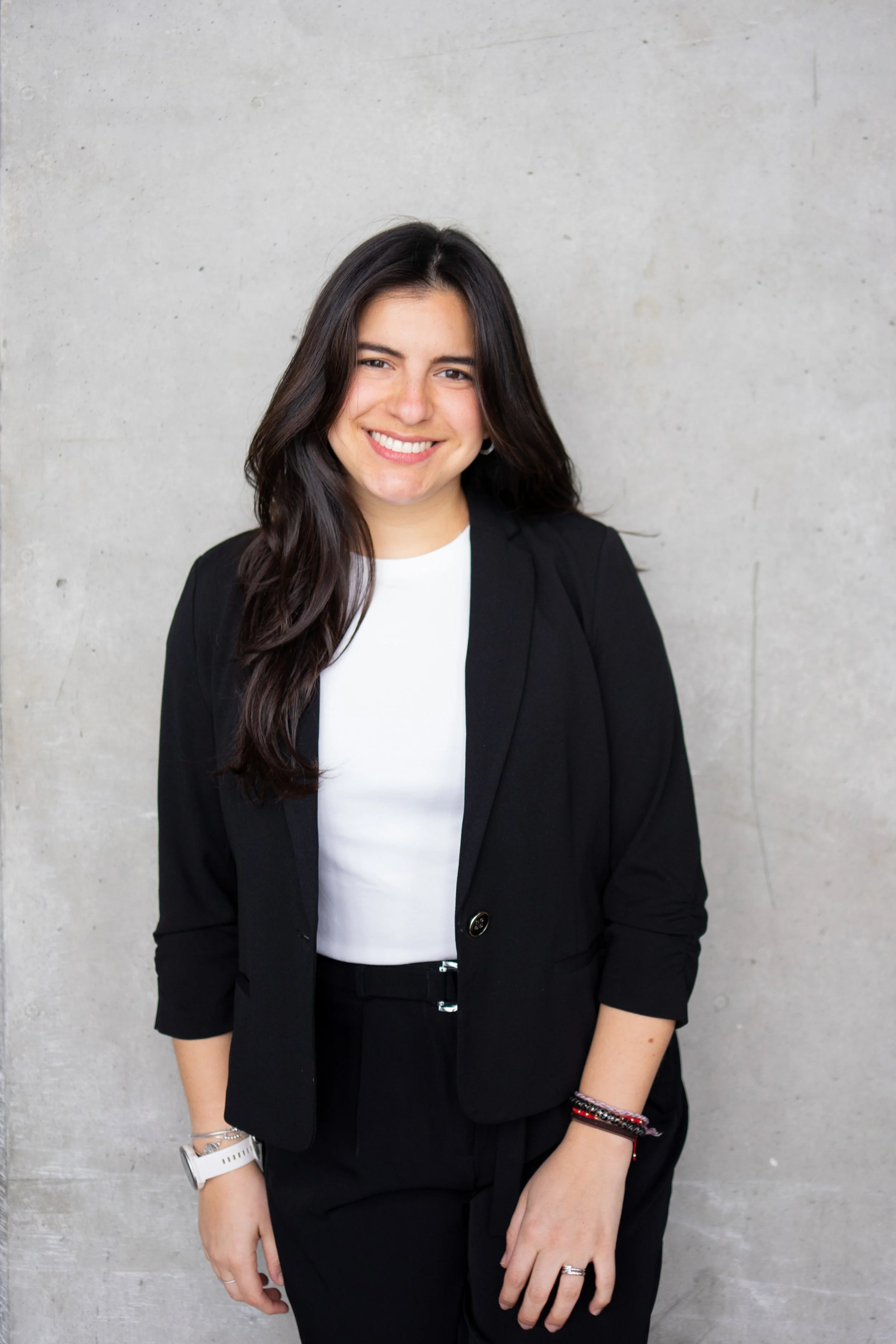A woman with long dark hair wearing a black blazer and black pants, smiling, standing against a plain concrete wall.