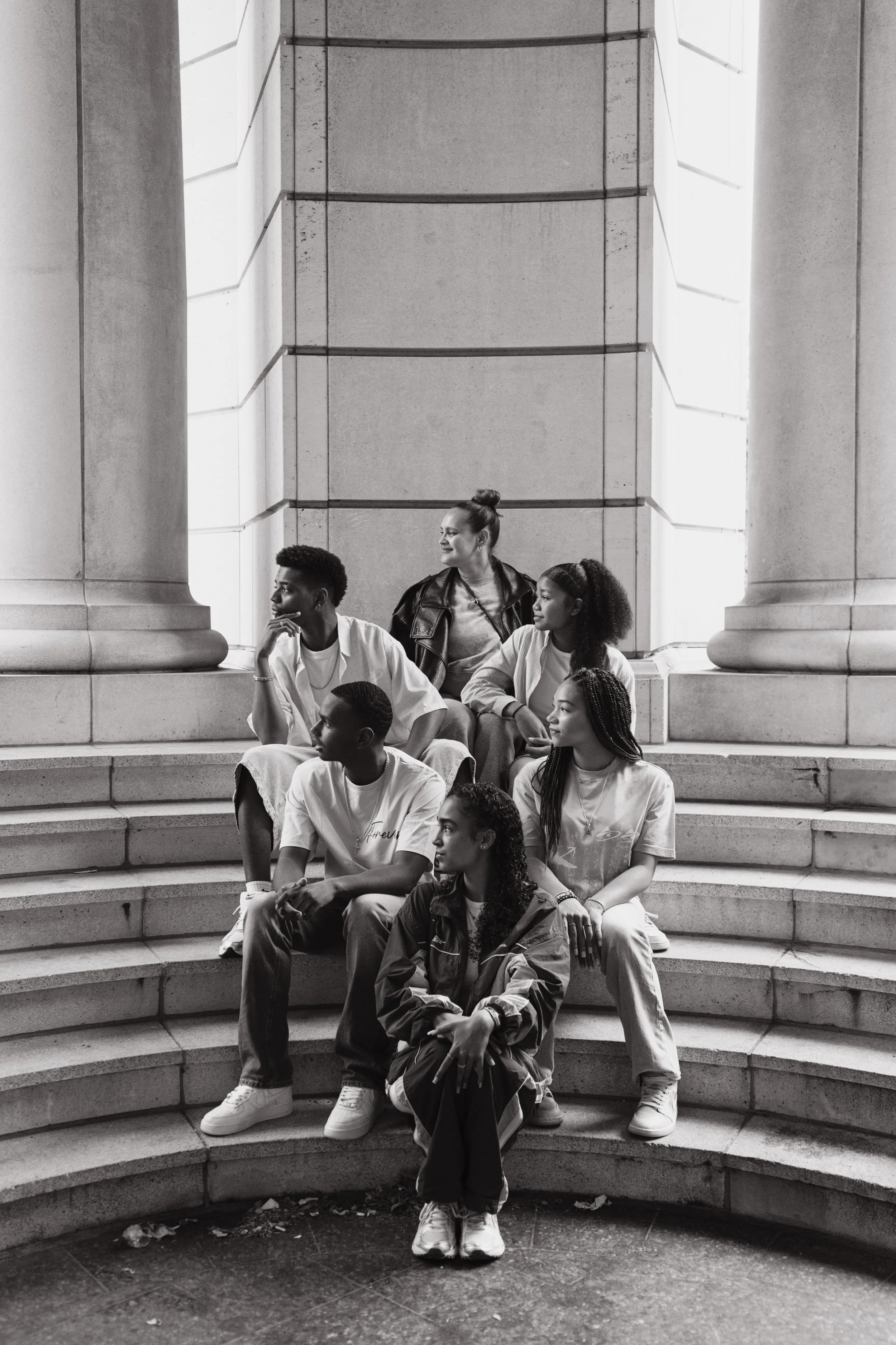 A group of young people sitting on the steps of a large building with a stone facade, some looking to the side and some looking down, in black and white.