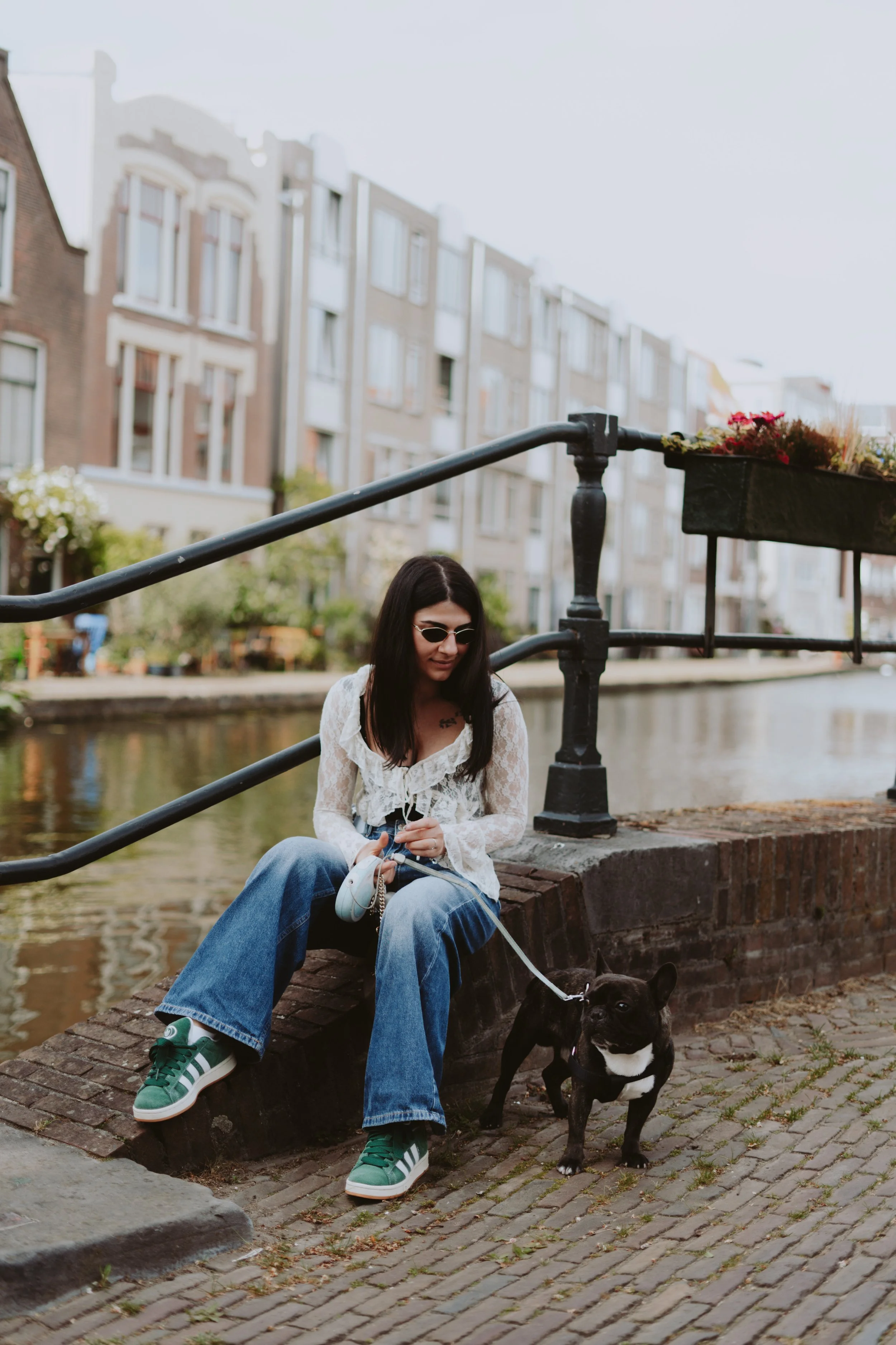 A woman sitting on a brick ledge beside a canal, holding a leash with a French Bulldog dog, wearing sunglasses, a white lace top, and baggy blue jeans, with city buildings in the background.