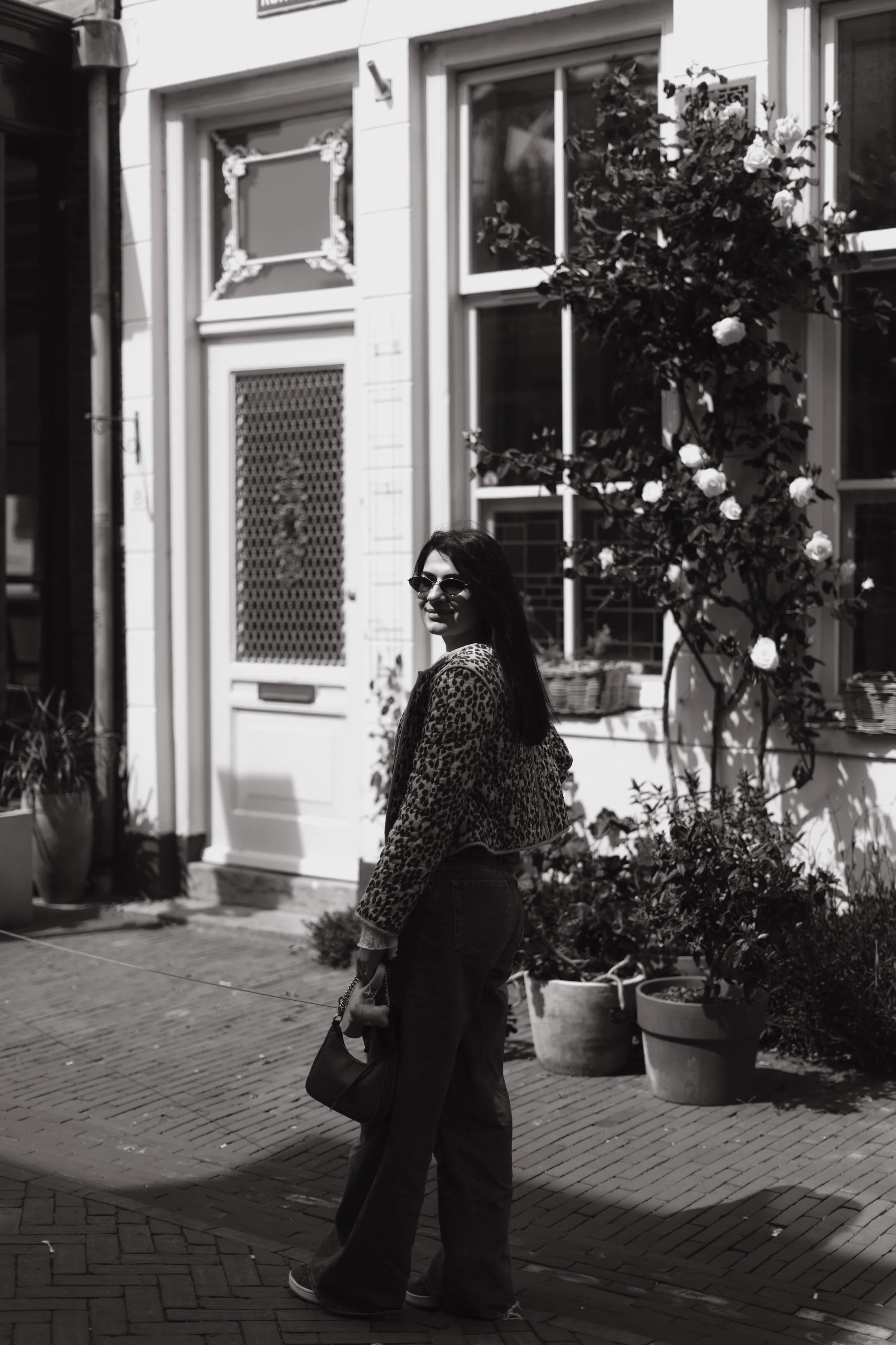 A woman with long hair wearing sunglasses, a leopard-print jacket, and dark pants, stands outside near a building with potted plants and a flowering tree, smiling and looking over her shoulder.