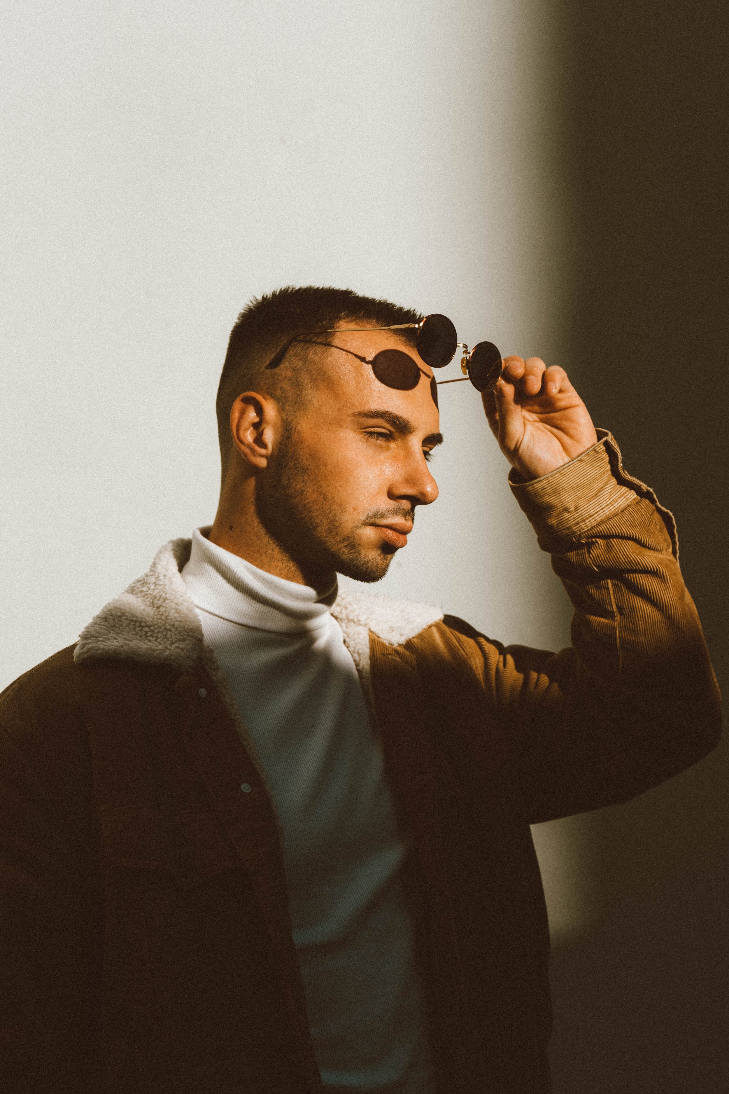 A young man with short dark hair and a beard is adjusting black sunglasses on his head. He is wearing a brown jacket with a fuzzy collar and a white turtleneck sweater. The background is plain with a gradient from light to dark.