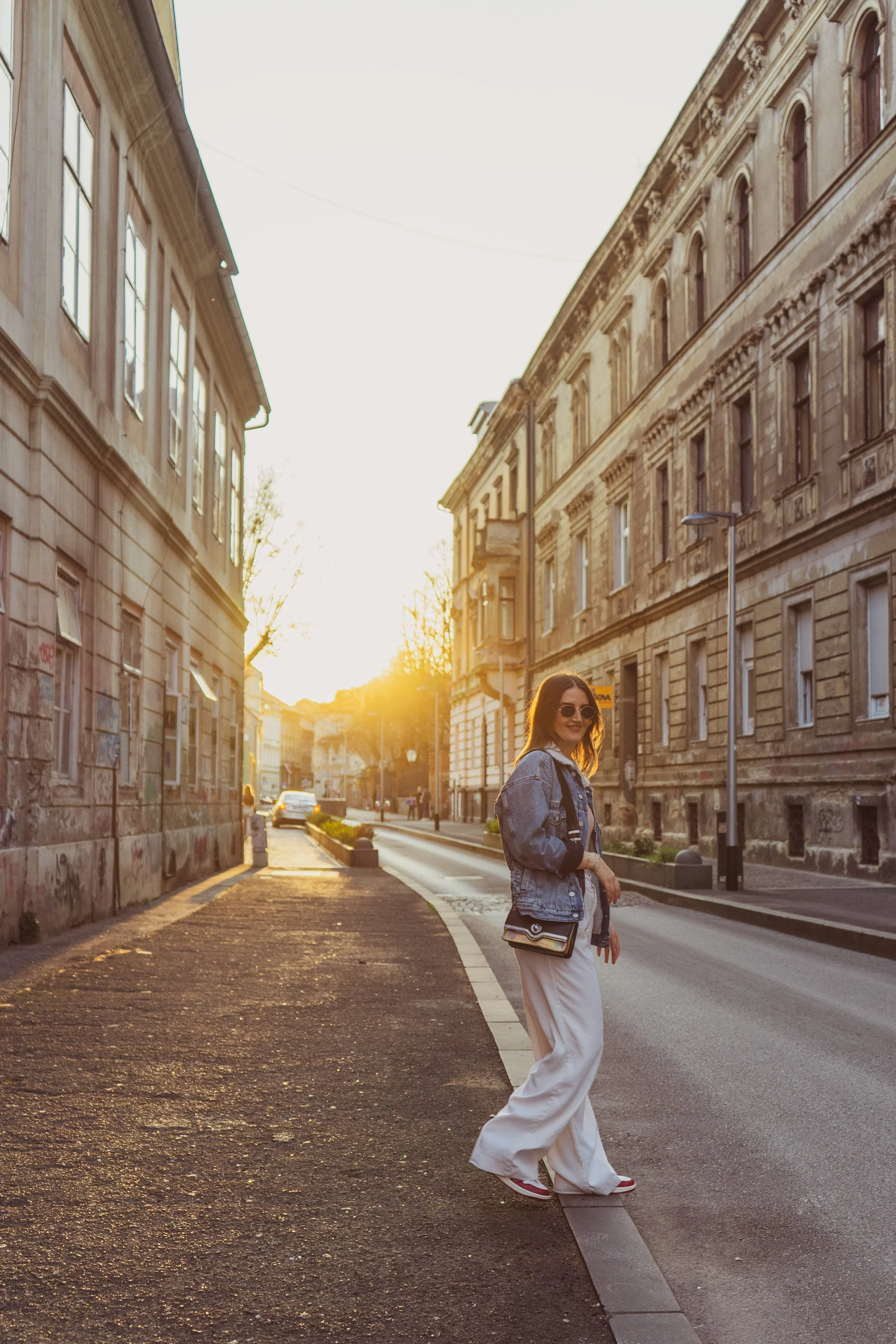 A young woman with sunglasses and a denim jacket walking on a city sidewalk during sunset, with old buildings on either side.