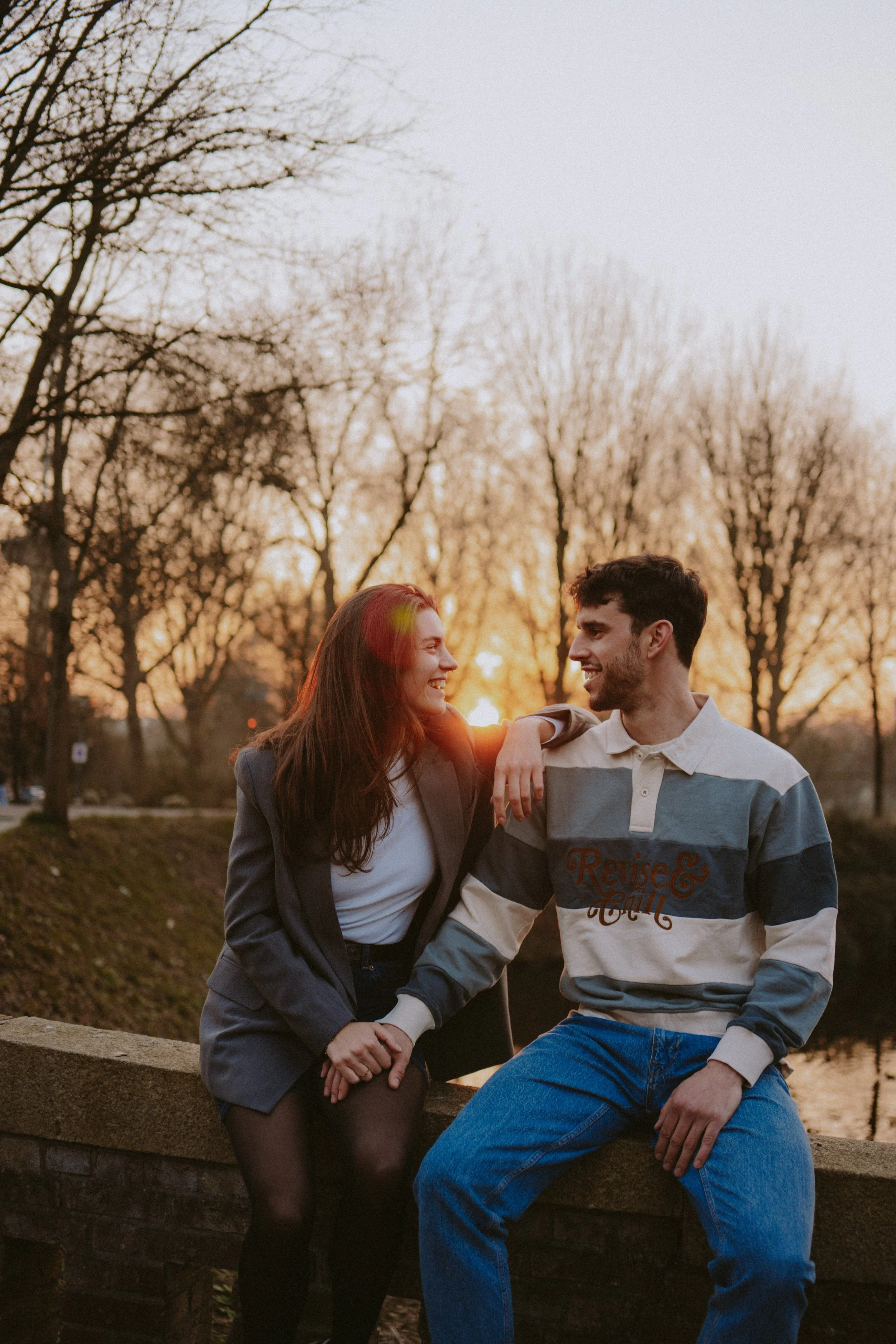 A young couple sitting on a brick ledge outdoors during sunset, holding hands and smiling at each other, with bare trees in the background.