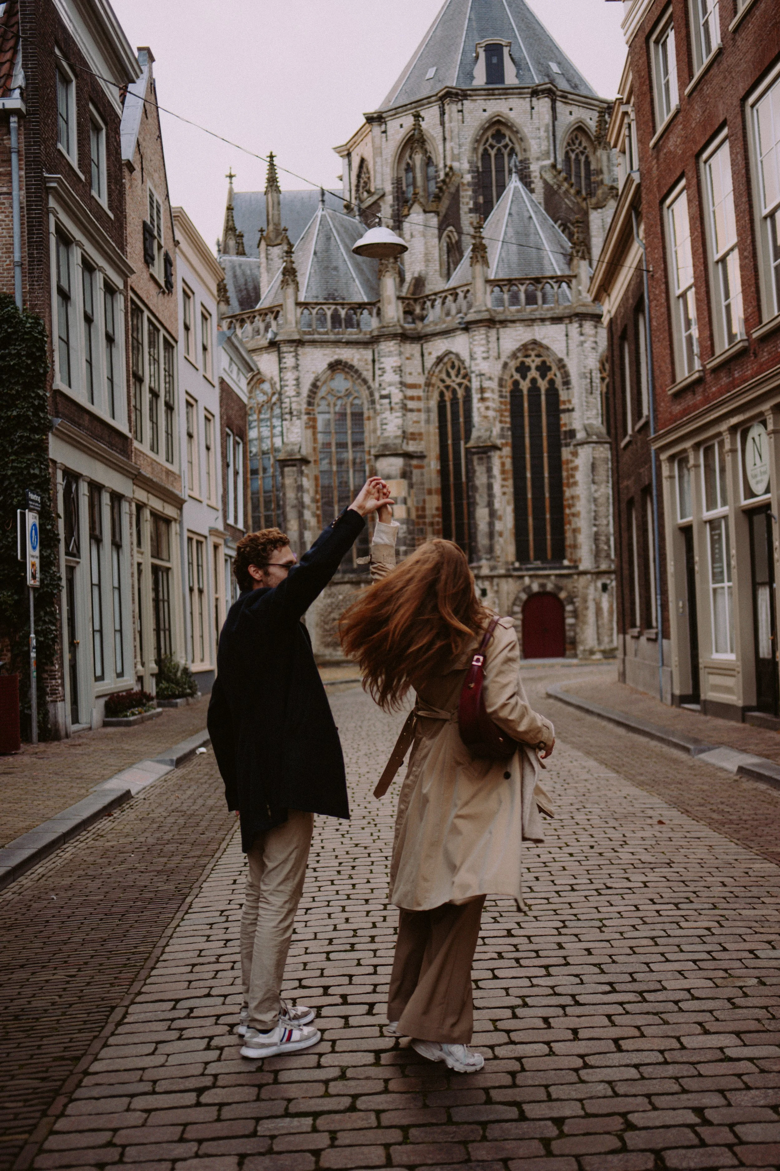 Two people dancing on a cobblestone street in front of a historic church. The man has brown curly hair, glasses, and is wearing a black coat with beige pants. The woman has long red hair, a beige trench coat, and a maroon bag. They are holding hands 