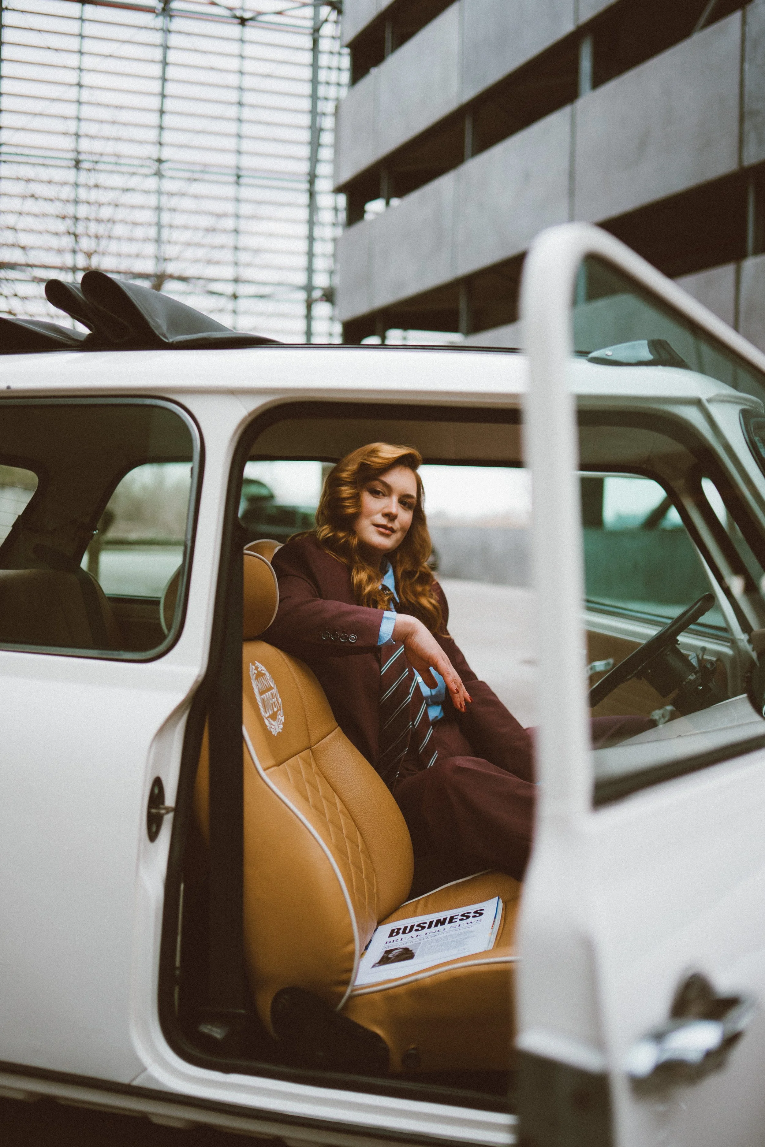 Editorial photography of a woman with brown hair sitting inside a white car with tan leather seats, holding a business magazine, with a modern building in the background.