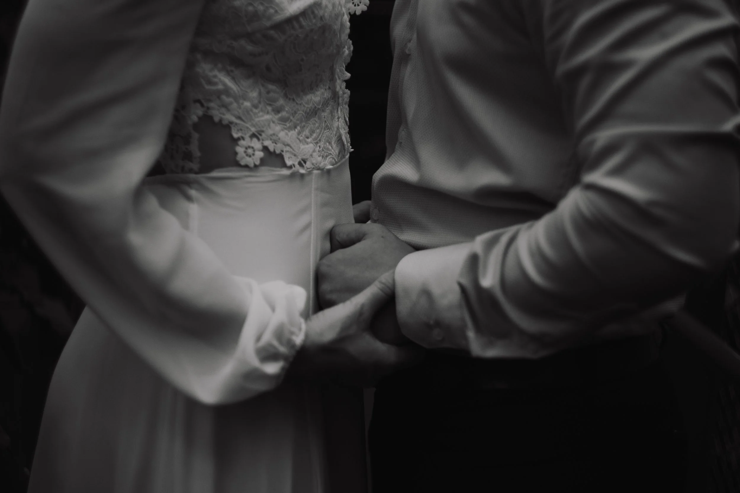 Close-up of a person dressed in formal white shirt holding the hand of a woman wearing a lace dress or top, with the image captured from above in black and white.