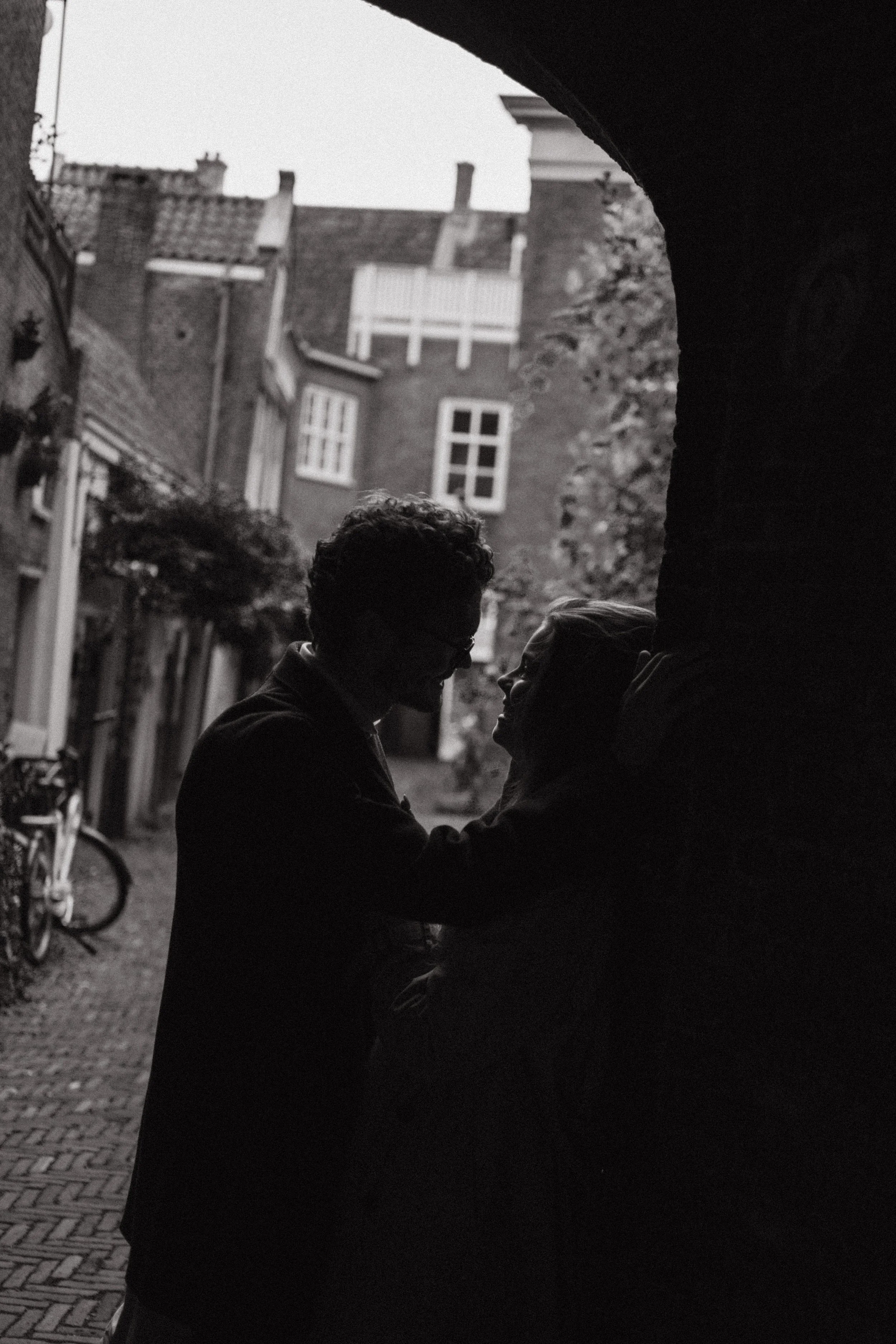 Two people facing each other, smiling and engaging in a conversation under an arched passageway with buildings and a bicycle visible in the background.