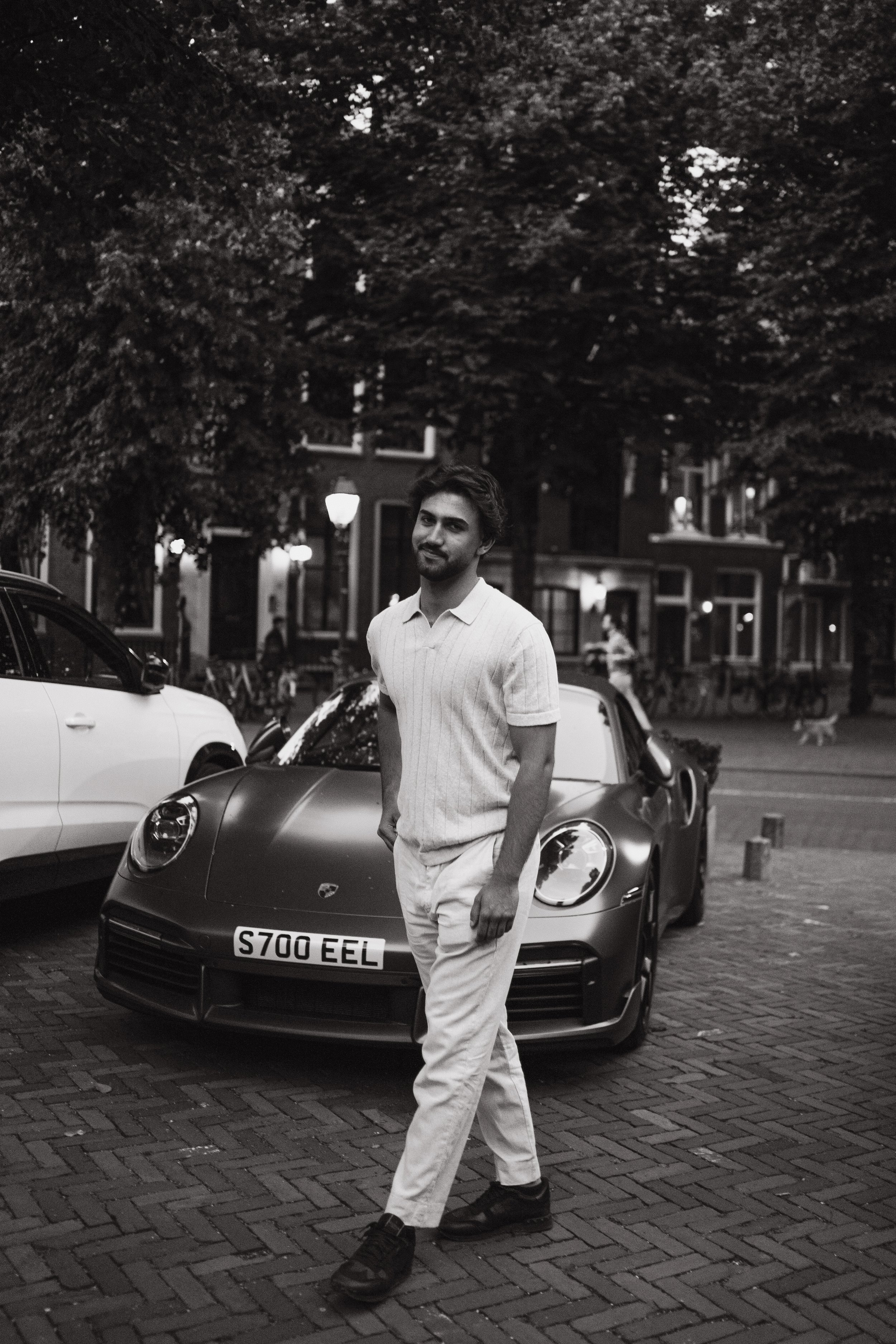 A young man with dark hair and a beard standing outdoors at night, wearing a white polo shirt and light-colored pants. He is standing in front of a front yard with parked cars and brick houses background, with trees illuminated above.