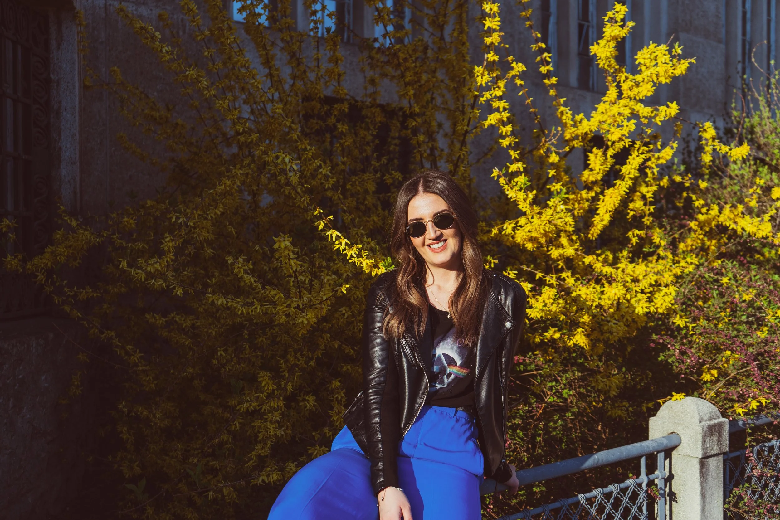 A woman with long brown hair wearing sunglasses, a black leather jacket, a graphic t-shirt, and blue pants, sitting on a bench in front of a yellow flowering shrub and stone wall.