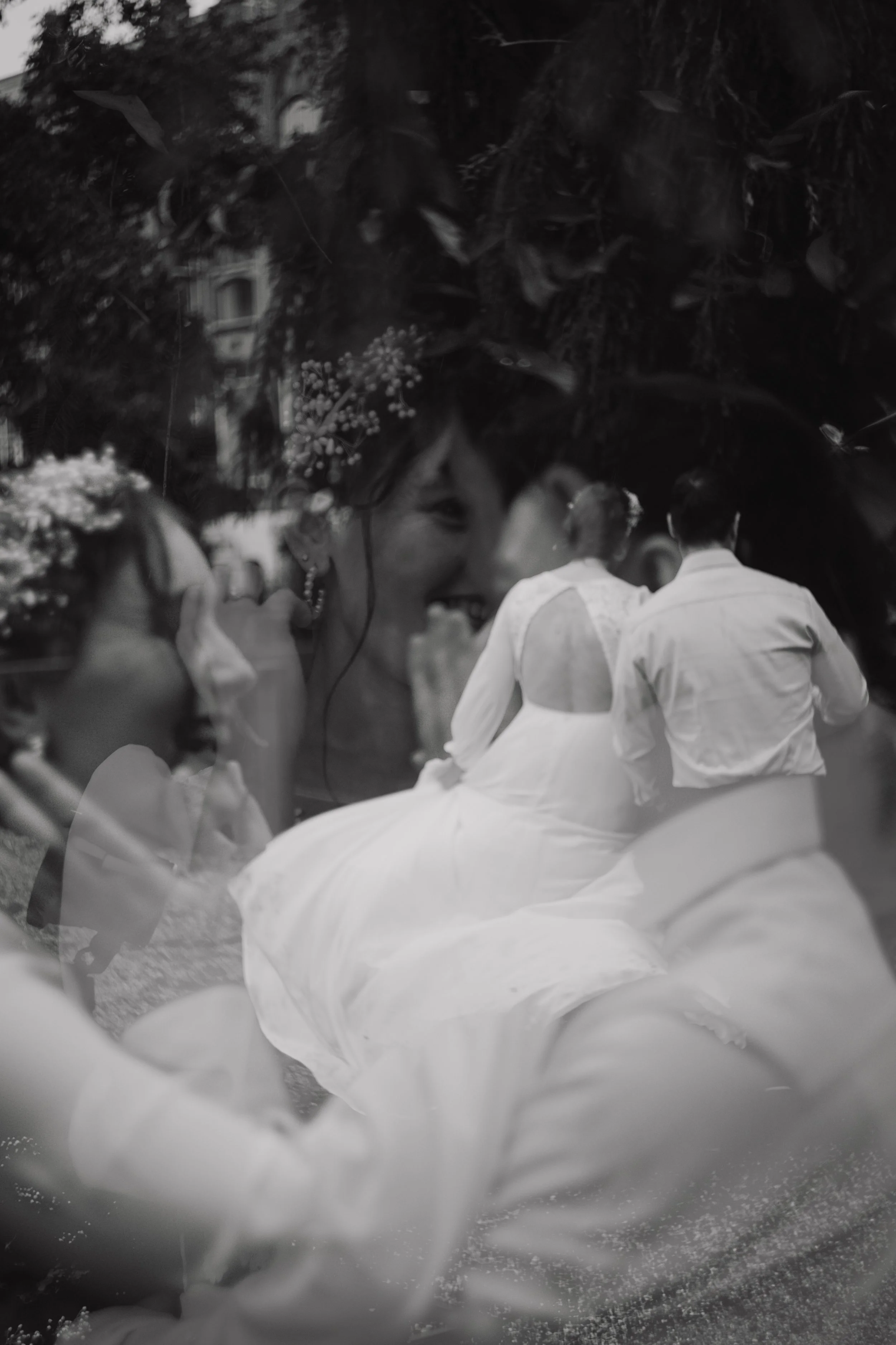 A black and white photograph depicting a romantic wedding scene outdoors with a bride and groom sitting on a bench surrounded by lush greenery, trees, and flowers, with an urban building in the background.