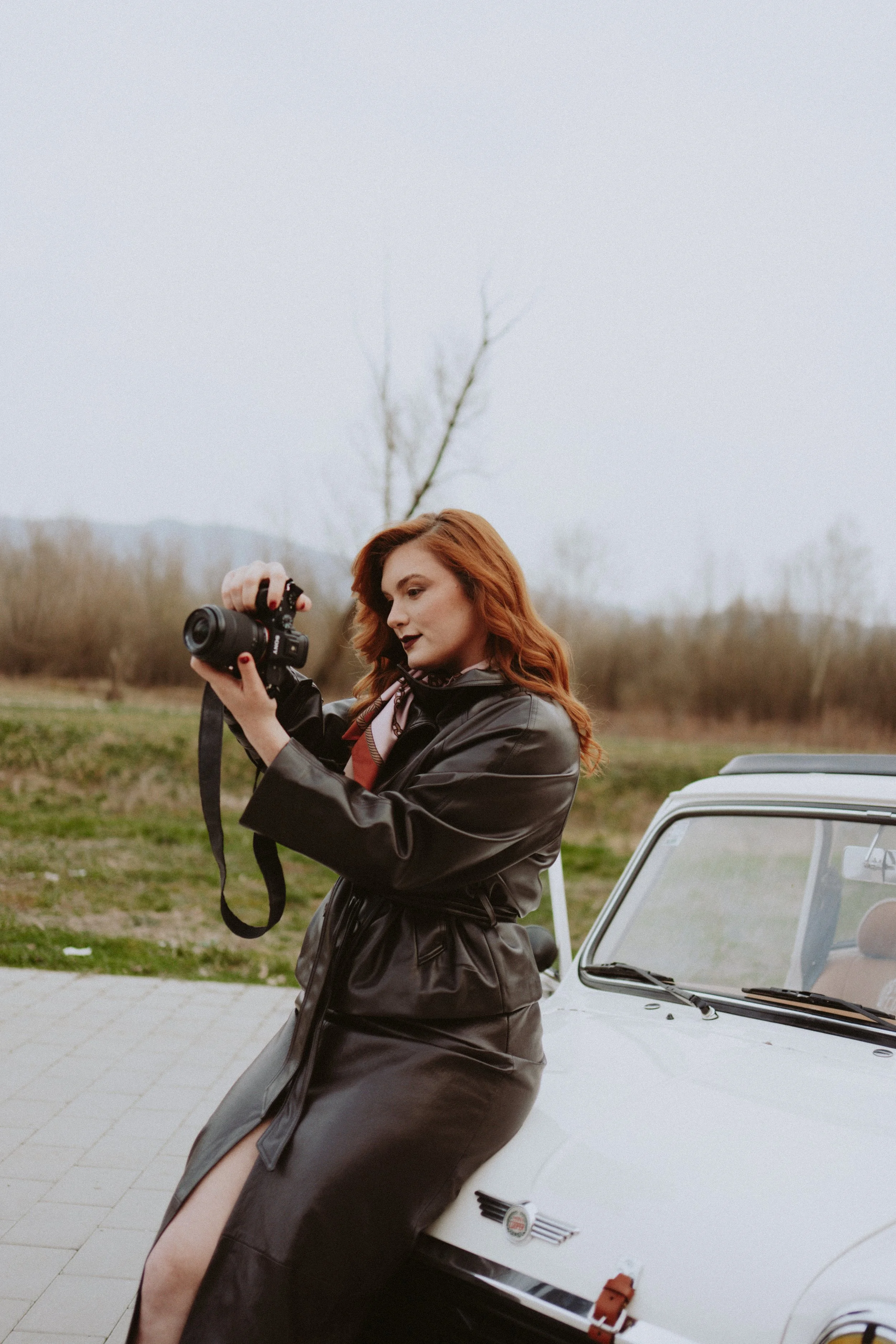 A young woman with red hair is standing outdoors next to a white vintage car, holding a camera and looking at the viewfinder.