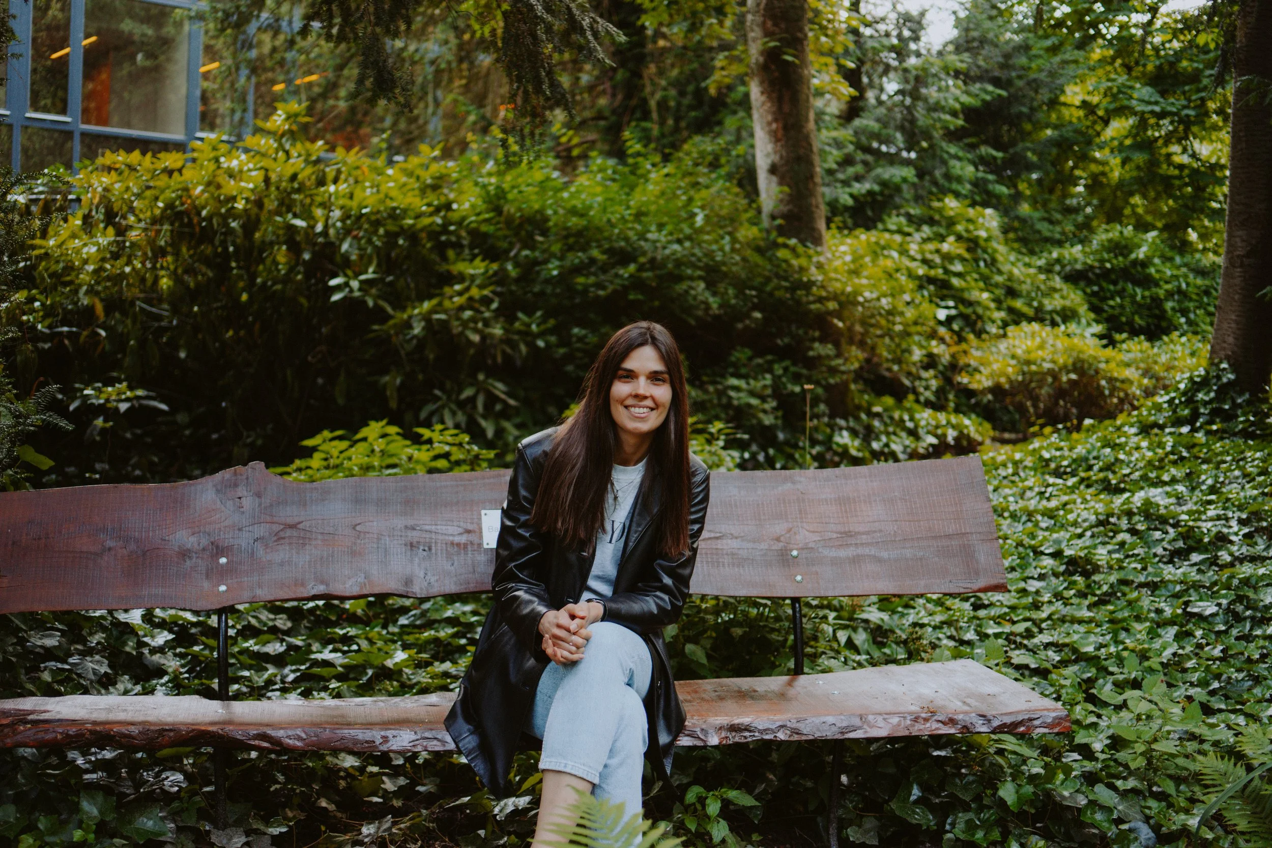 A young woman with long dark hair, smiling, seated on a wooden park bench with a green forest background, wearing a black jacket and light-colored pants.