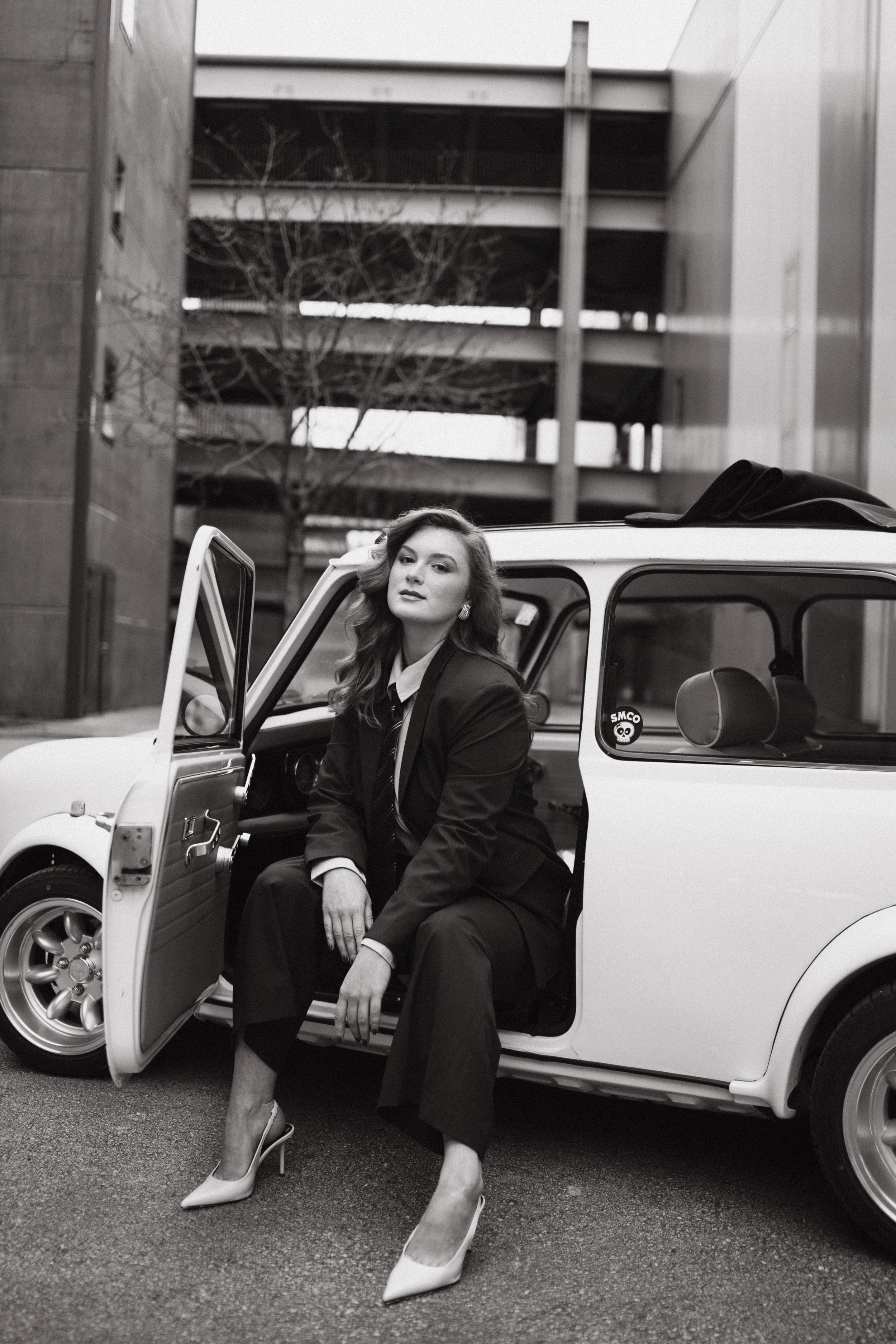 Timeless photography of  woman in a suit sitting on the open door step of a small convertible car parked between two buildings. The woman has wavy hair and is wearing high heels.