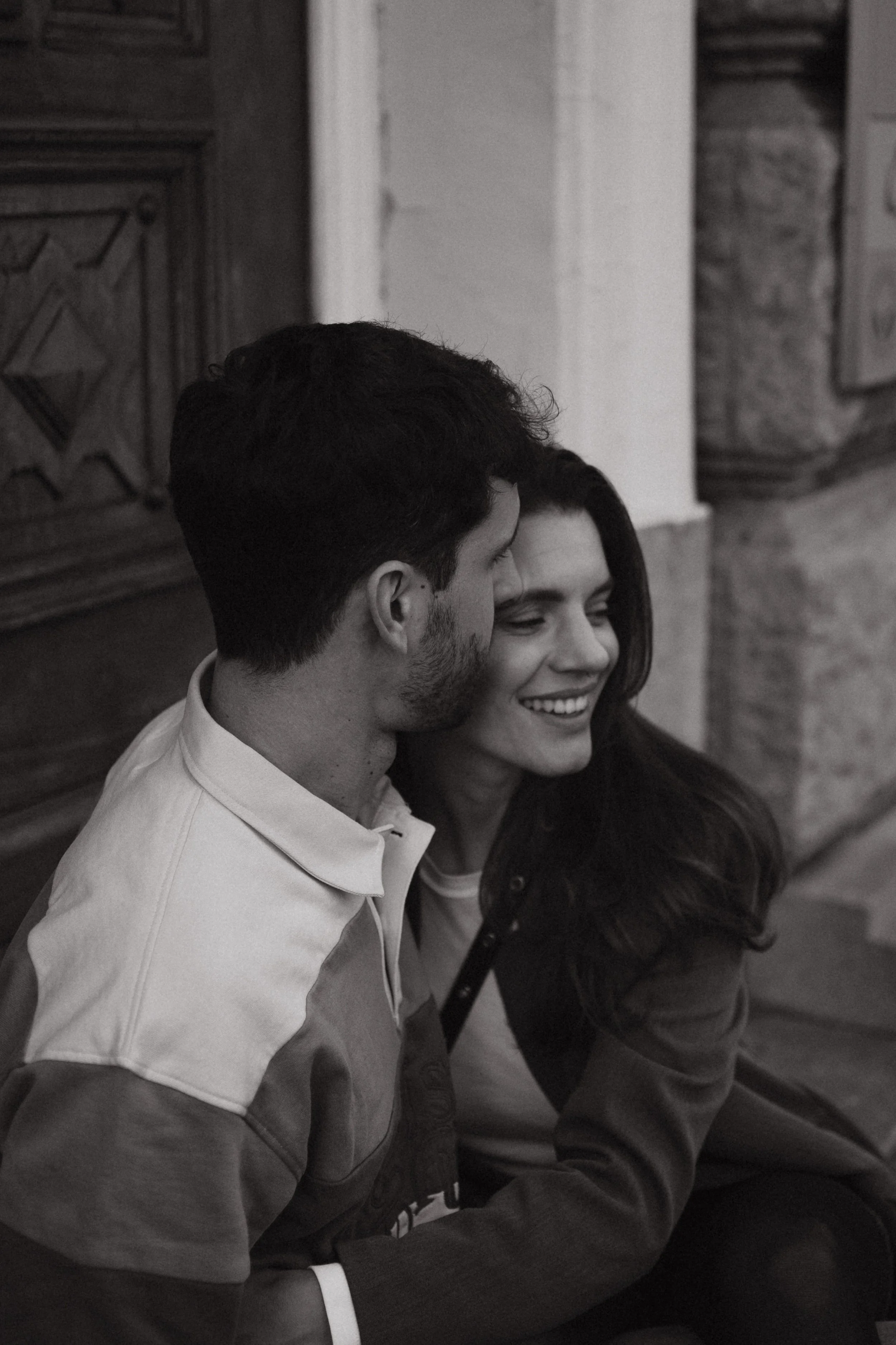 A black-and-white photo of three people sitting together, close to each other, smiling, against a background of wood and stone walls.