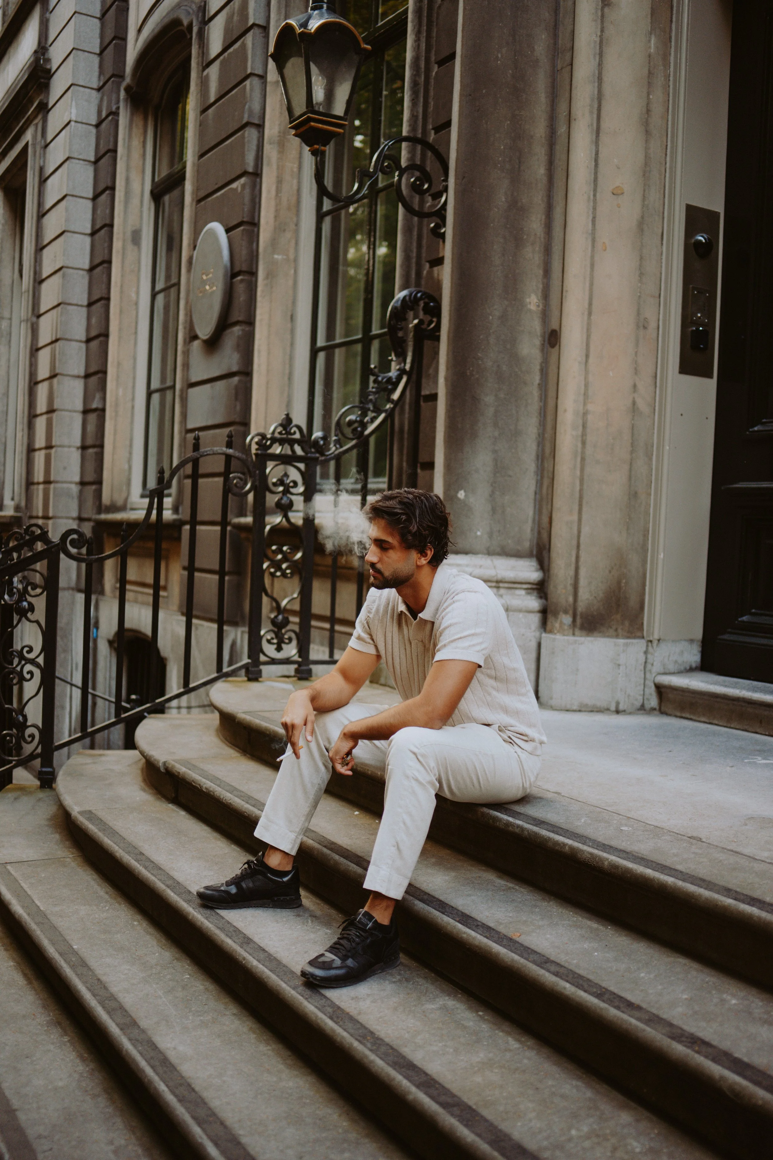 A young man with dark hair and a beard sitting on stone steps outside a building, wearing a beige short-sleeve shirt and light-colored pants, with a cigarette in his mouth, and black sneakers, surrounded by ornate black iron railings.
