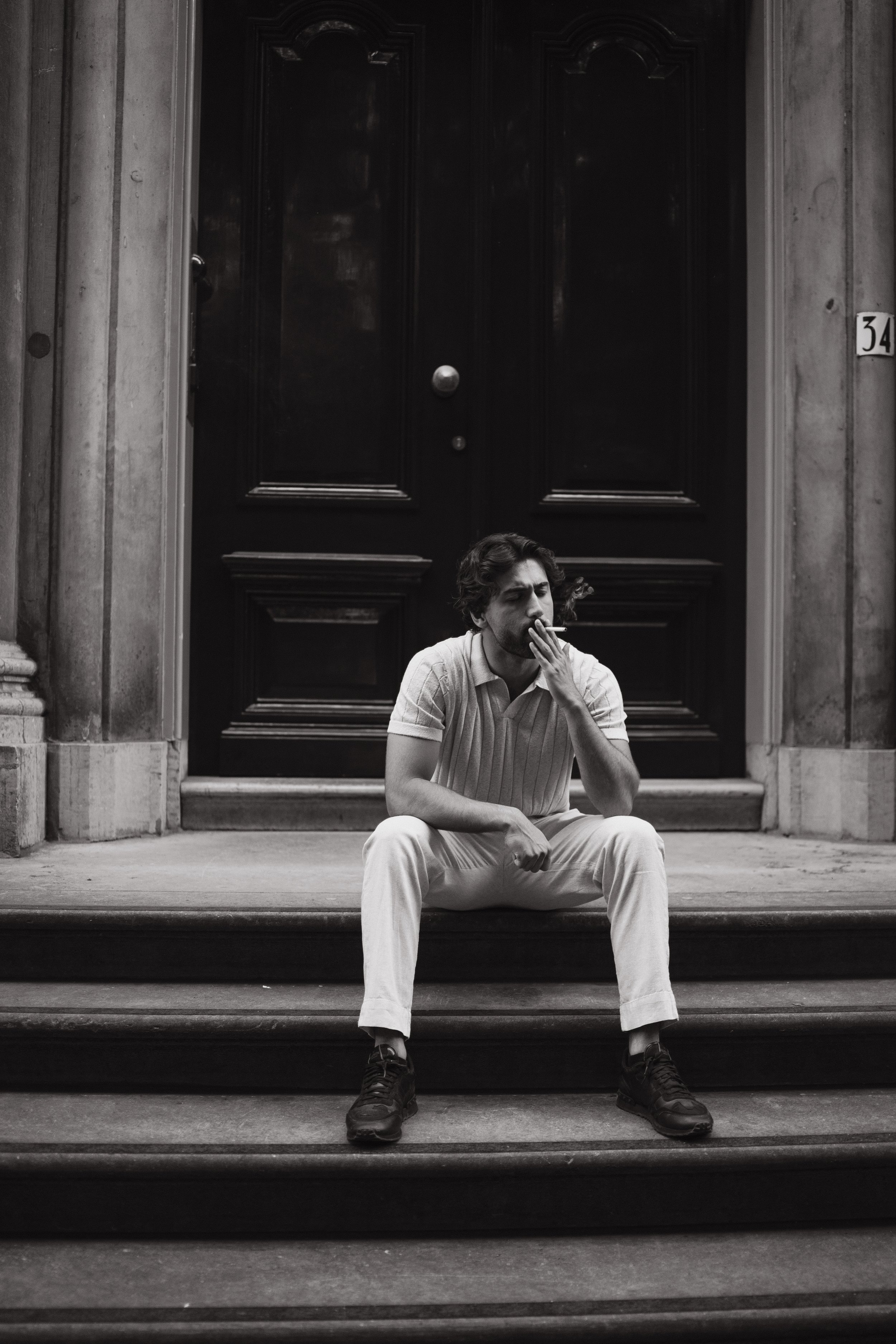 A man with curly hair wearing a light-colored, short-sleeved shirt and light-colored pants is sitting on the steps in front of a large, dark wooden door. He is smoking a cigarette and resting his left arm on his thigh.