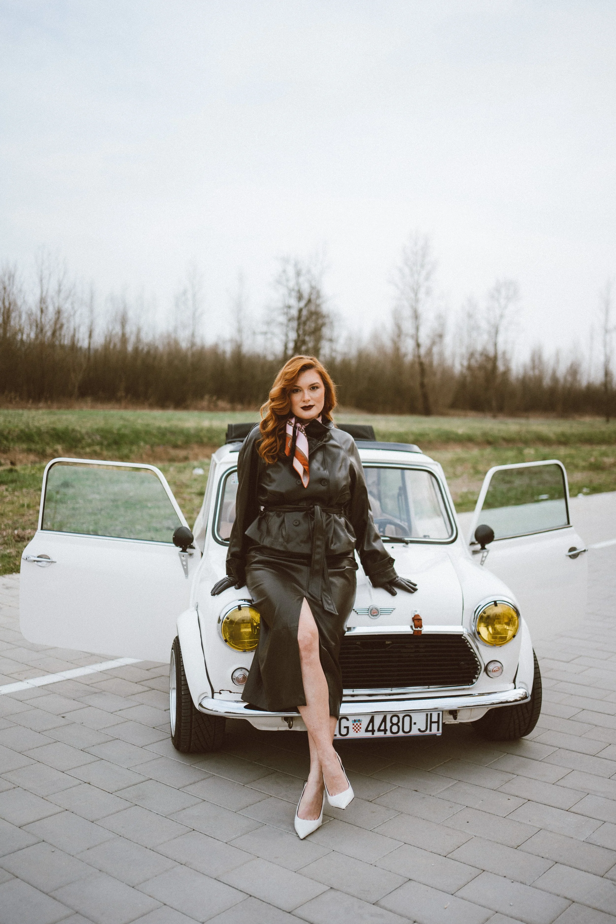 A woman with red hair in a black leather trench coat and white high heels poses in front of a vintage white car with open doors, parked on gray paving stones in an outdoor setting with trees and overcast sky in the background.