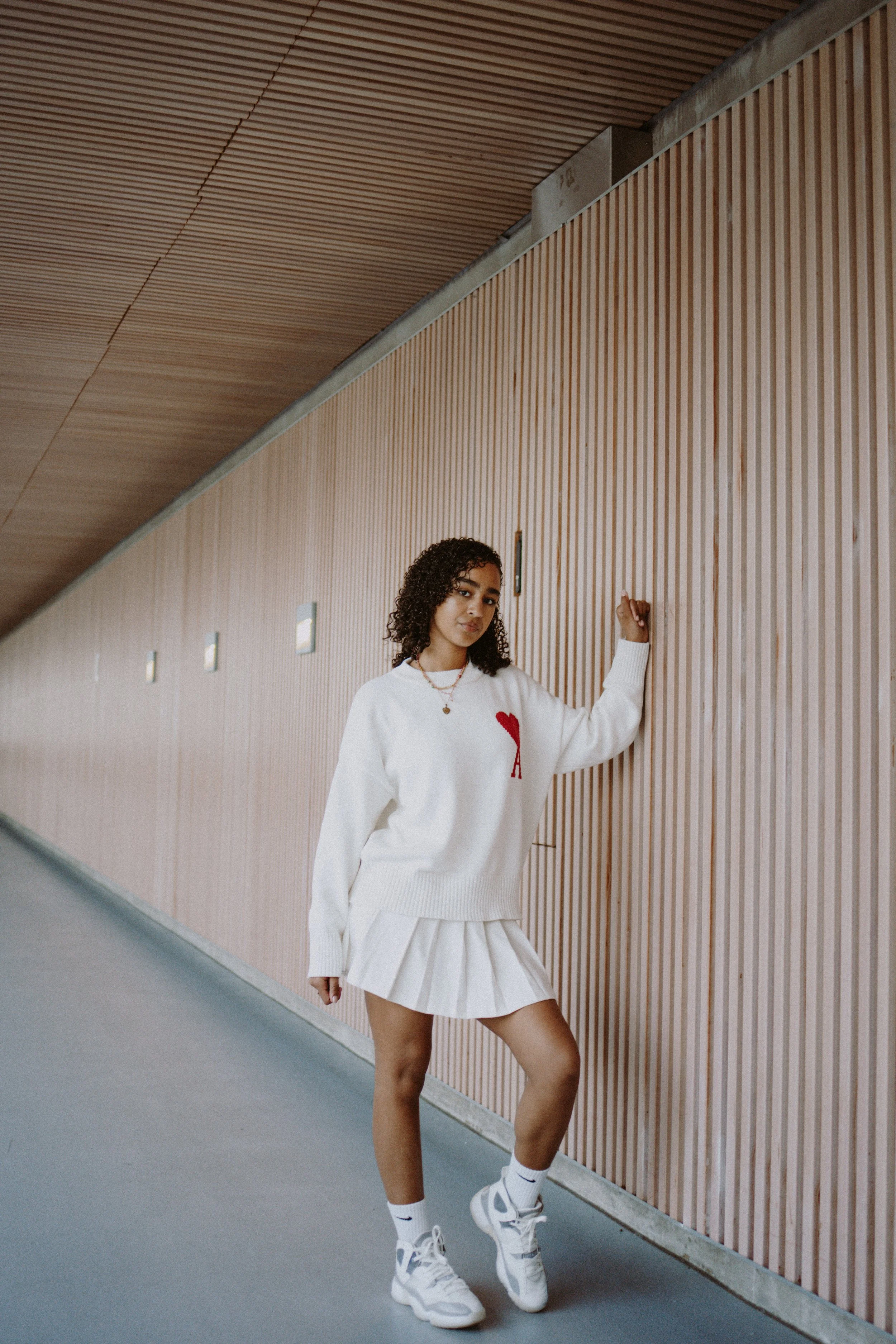 A young woman with curly hair standing next to a wooden paneled wall, wearing a white sweatshirt with a red heart and the letter 'A', a white pleated skirt, white Nike sneakers, and white socks, posing with her right hand on the wall.