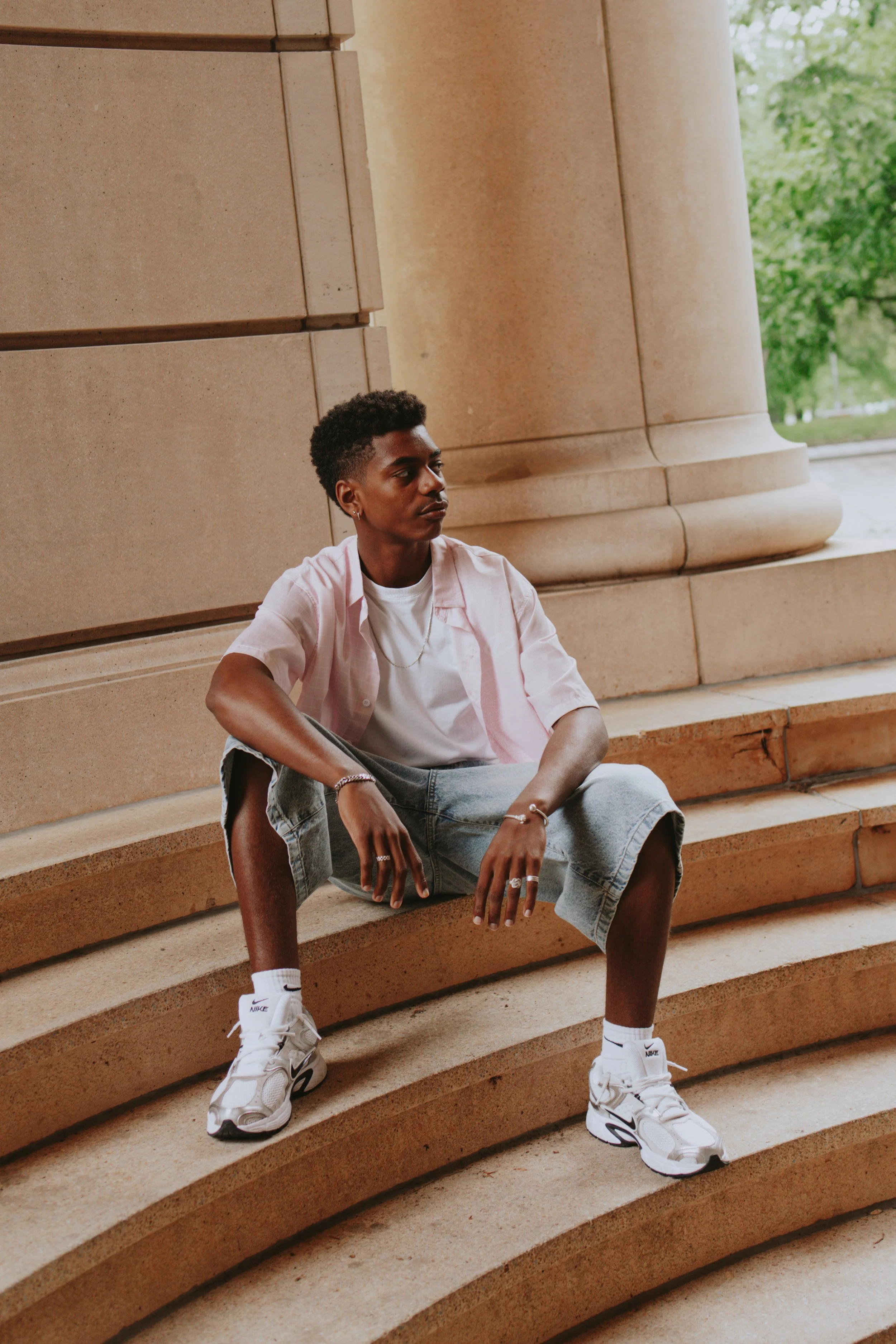 A young person with short curly hair sitting on stone steps outside in front of a building with large columns and greenery in the background. They are wearing a light pink shirt, white T-shirt, ripped jeans, white Nike sneakers, and accessories, with